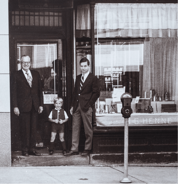 Black and White Photo of an Older Man and Middle Aged Man, both in Suits, and a Young Boy in front of the Henne Storefront