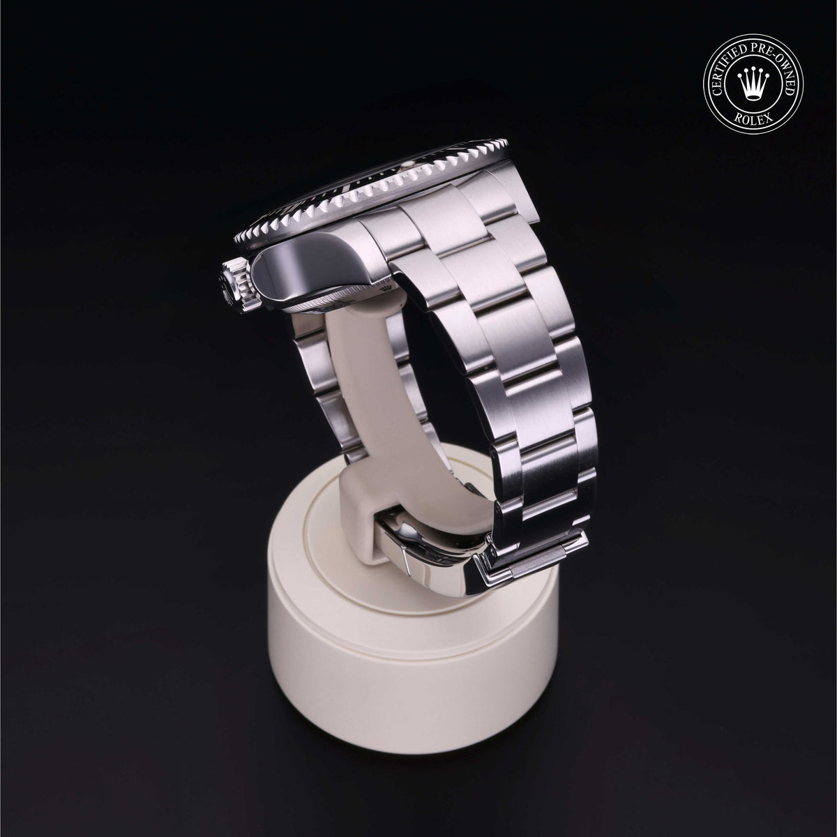 Side view of a stainless steel luxury watch with a metal link bracelet displayed on a beige stand against a black background, featuring a Rolex Certified Pre-Owned logo in the corner.