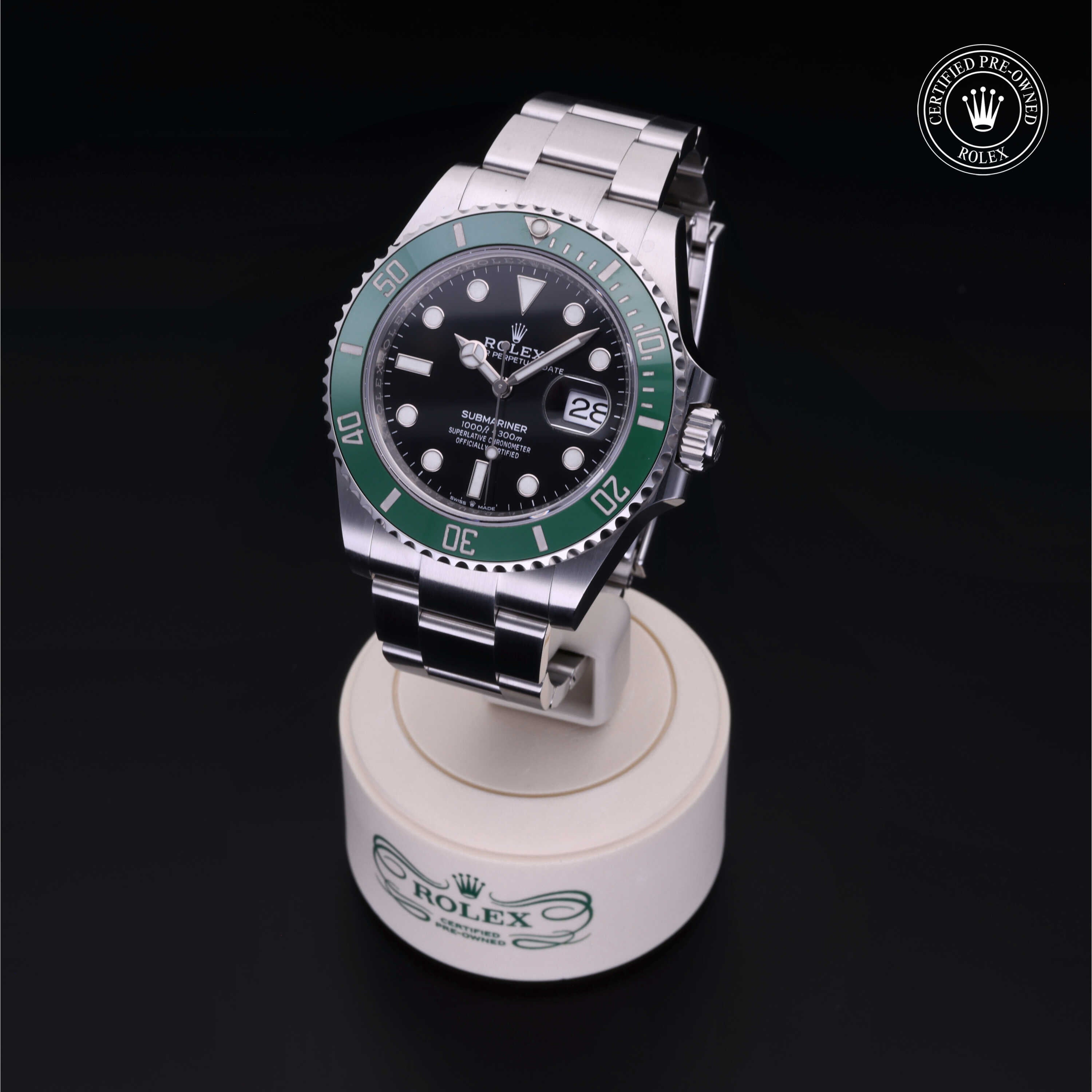 Rolex stainless steel watch with a green bezel and black dial displayed on a branded stand, featuring a date window and Certified Pre-Owned Rolex logo.