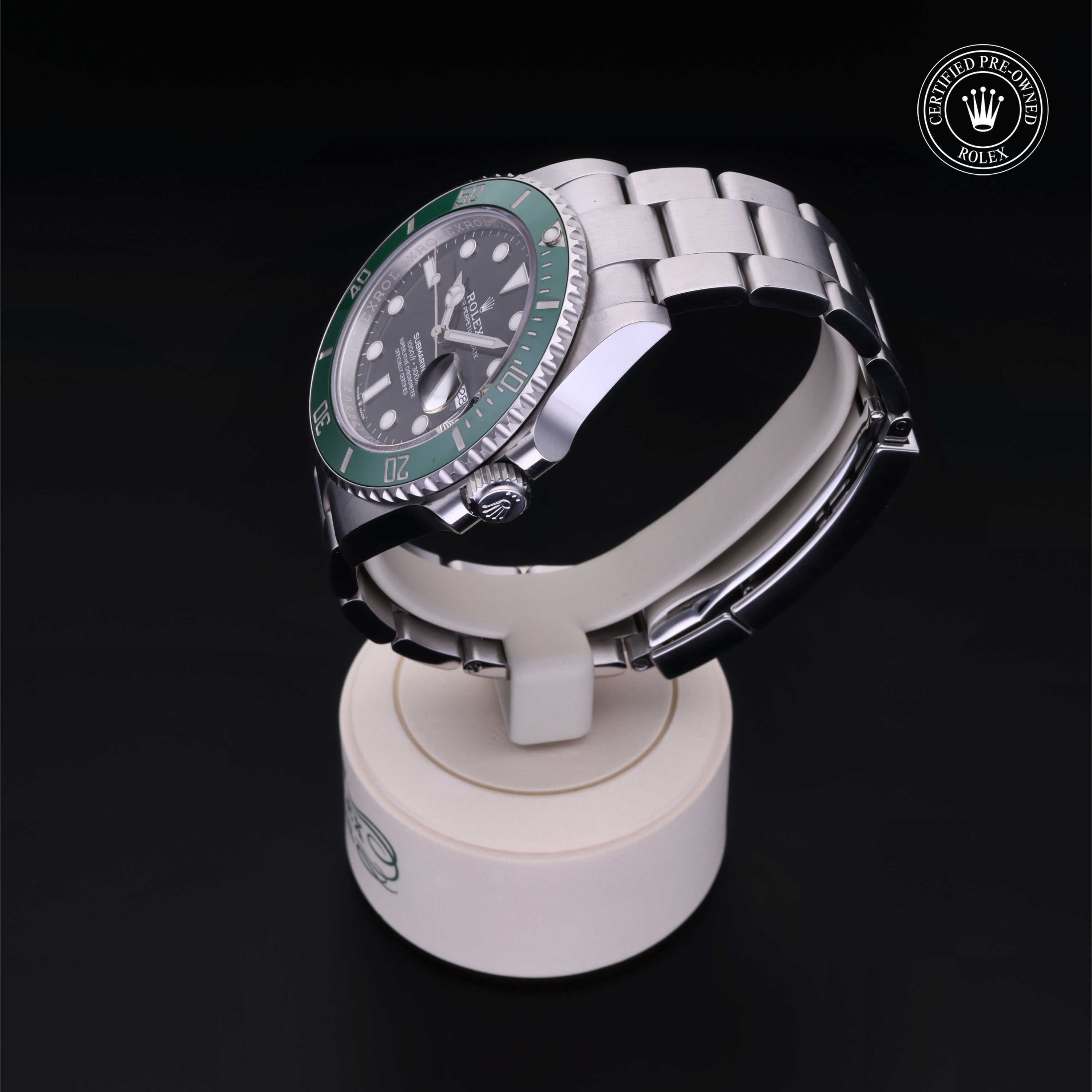 Close-up of a Rolex luxury watch with a green bezel and silver metal bracelet displayed on a white stand against a dark background, featuring a certified pre-owned emblem.