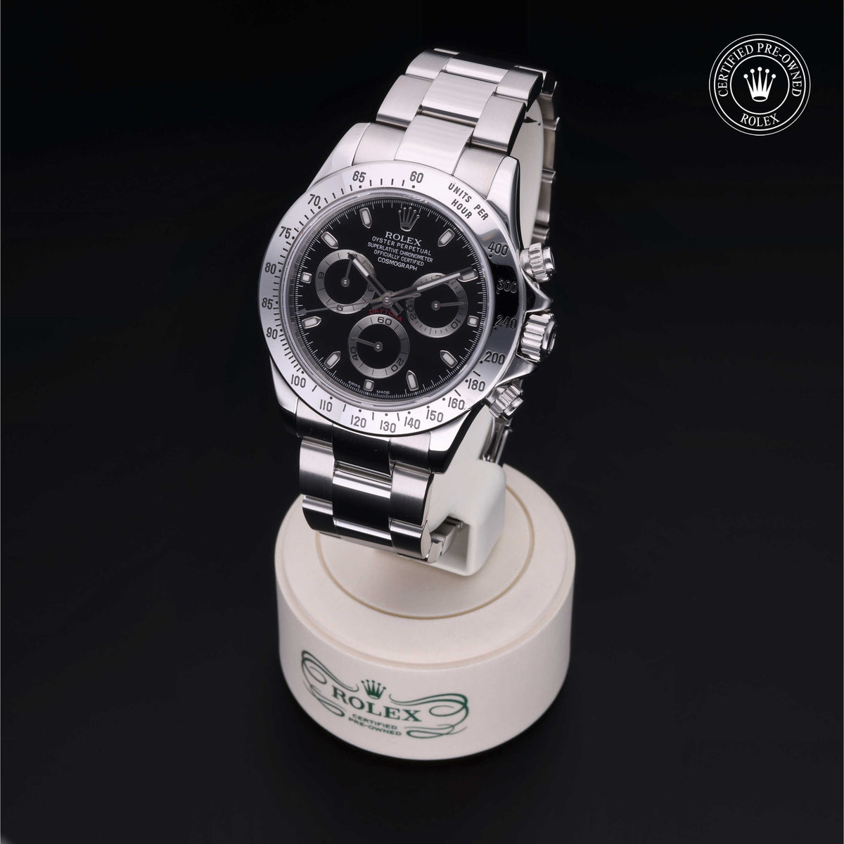 A luxury Rolex stainless steel watch with a black dial and chronograph features displayed on a branded white stand, marked as certified pre-owned.