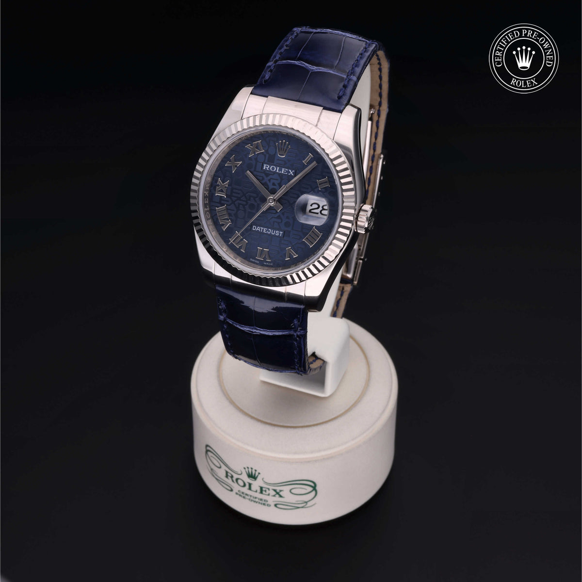 Close-up of a luxury Rolex wristwatch with a blue leather strap and Roman numeral markers, displayed on a white stand with Rolex branding and a certified pre-owned emblem.