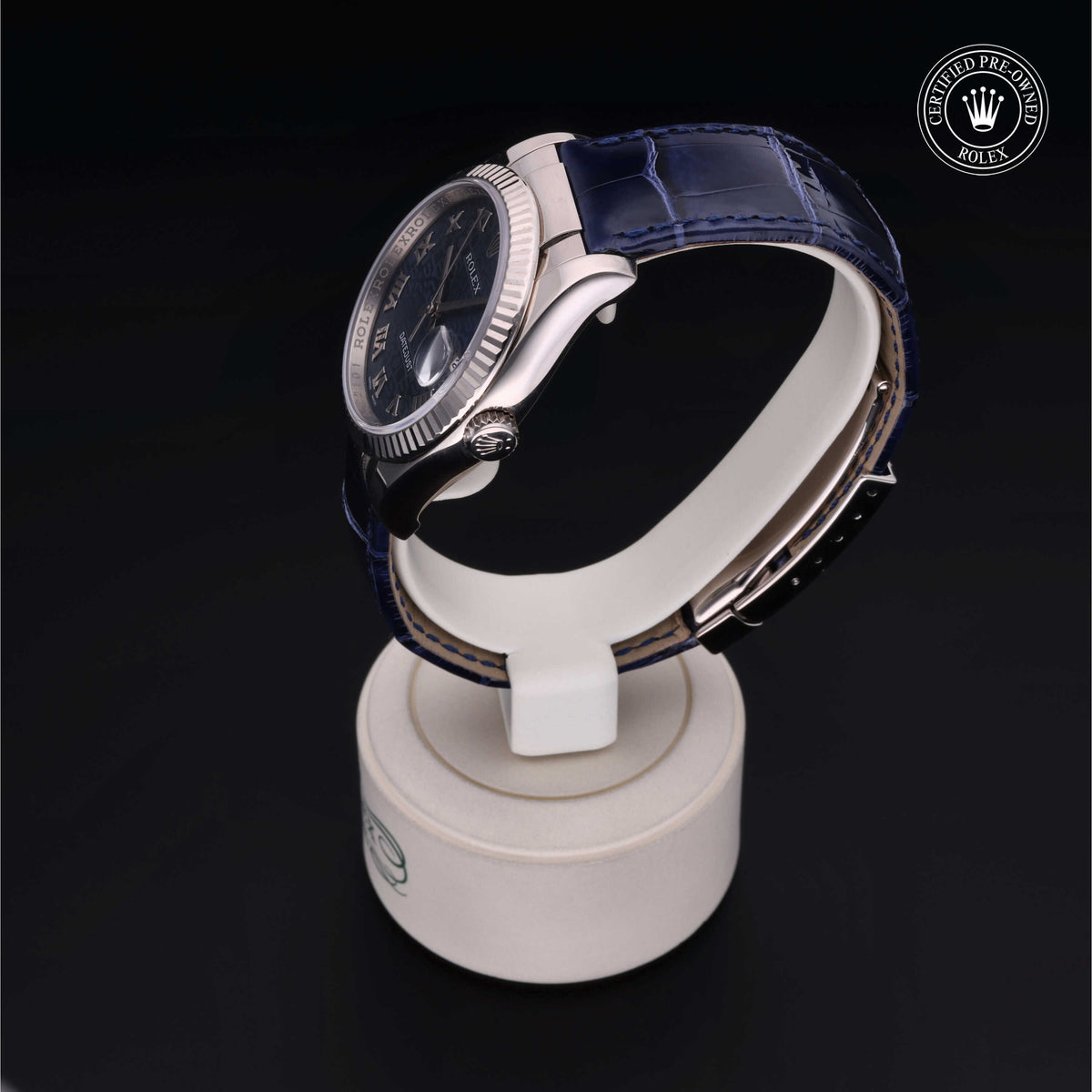Side view of a luxury Rolex watch with a dark blue leather strap displayed on a white stand against a black background, featuring a certified pre-owned logo.