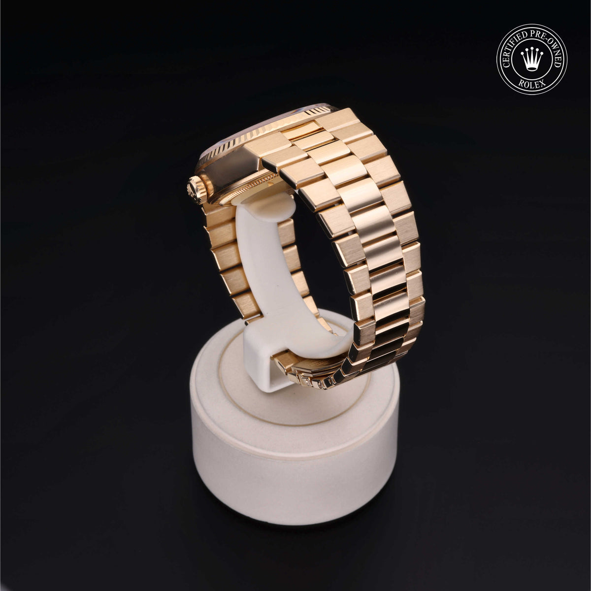 Close-up of a luxury gold Rolex watch band displayed on a white stand against a black background, featuring a certified pre-owned Rolex logo in the corner.