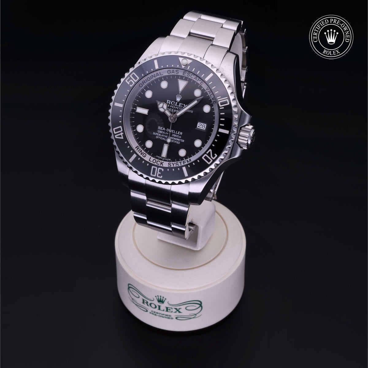 Rolex stainless steel luxury watch displayed on a branded stand against a black background, featuring a black dial and bezel with luminous markers and date window.