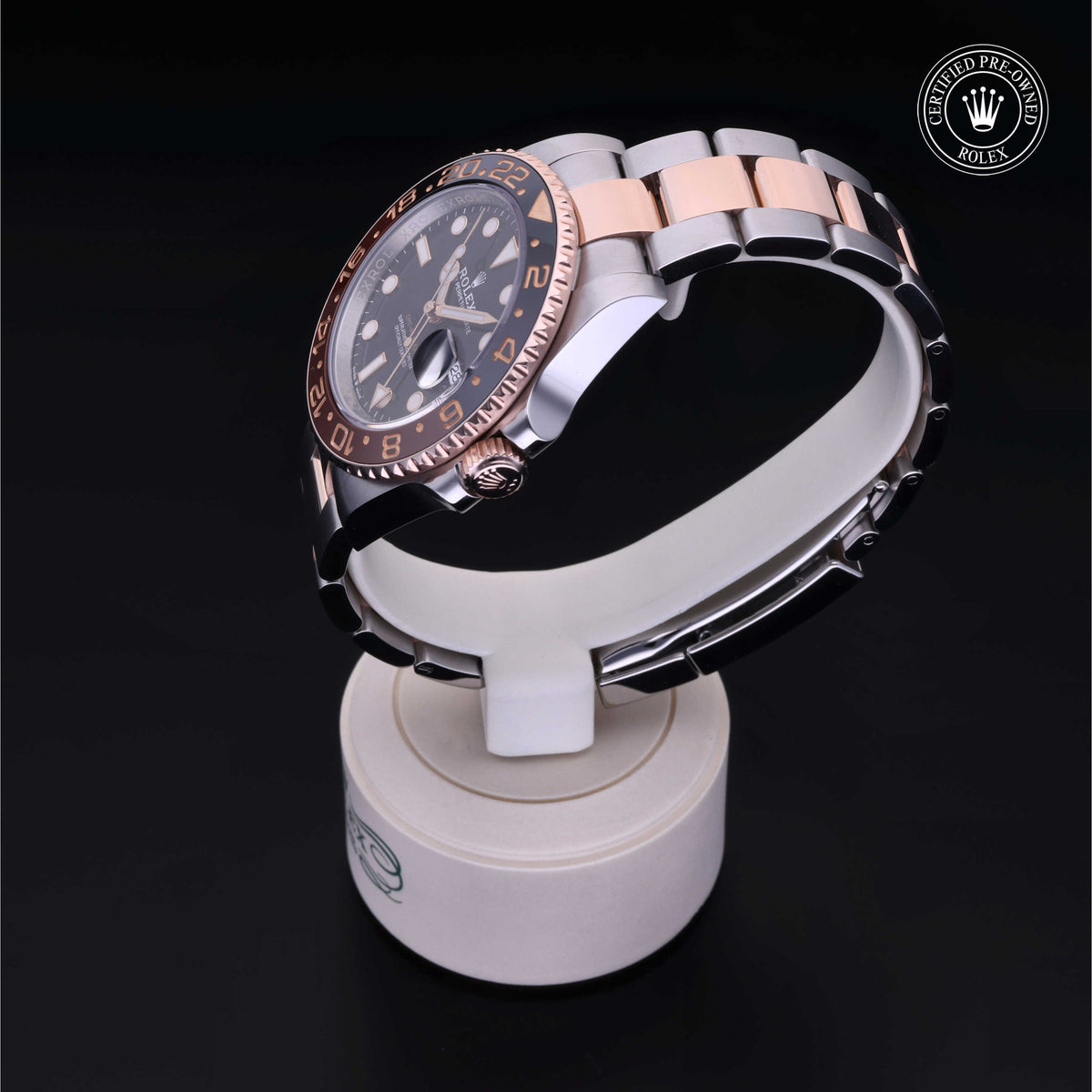 Two-tone luxury Rolex watch with a black dial and rose gold accents displayed on a white stand, highlighting its polished metal bracelet and certified pre-owned status.