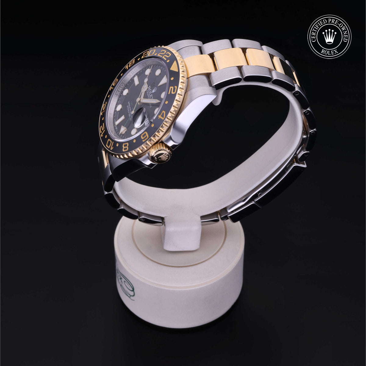 Two-tone Rolex wristwatch with a black and gold bezel, silver and gold metal bracelet, displayed on a white stand with a Certified Pre-Owned Rolex logo on a black background.