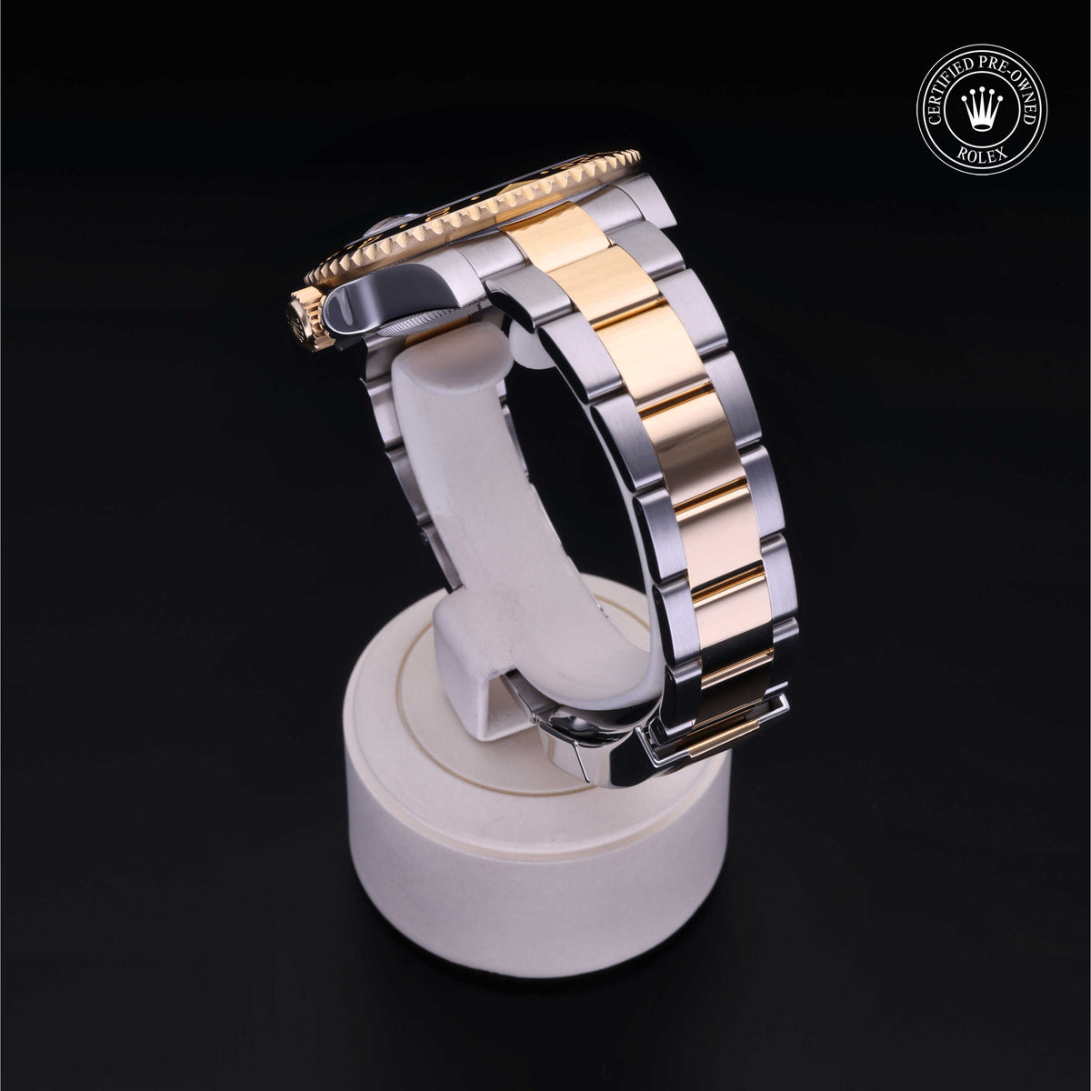 Two-tone luxury watch band in gold and silver displayed on a white stand against a black background, featuring a Rolex certified pre-owned logo.