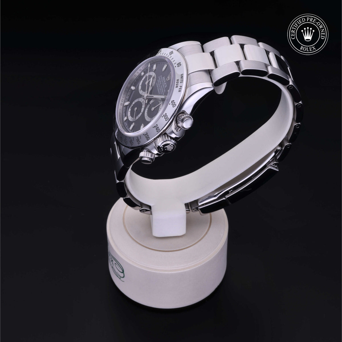 Silver Rolex chronograph luxury watch with black dial and stainless steel bracelet displayed on a white stand against a black background, featuring a certified pre-owned Rolex logo.
