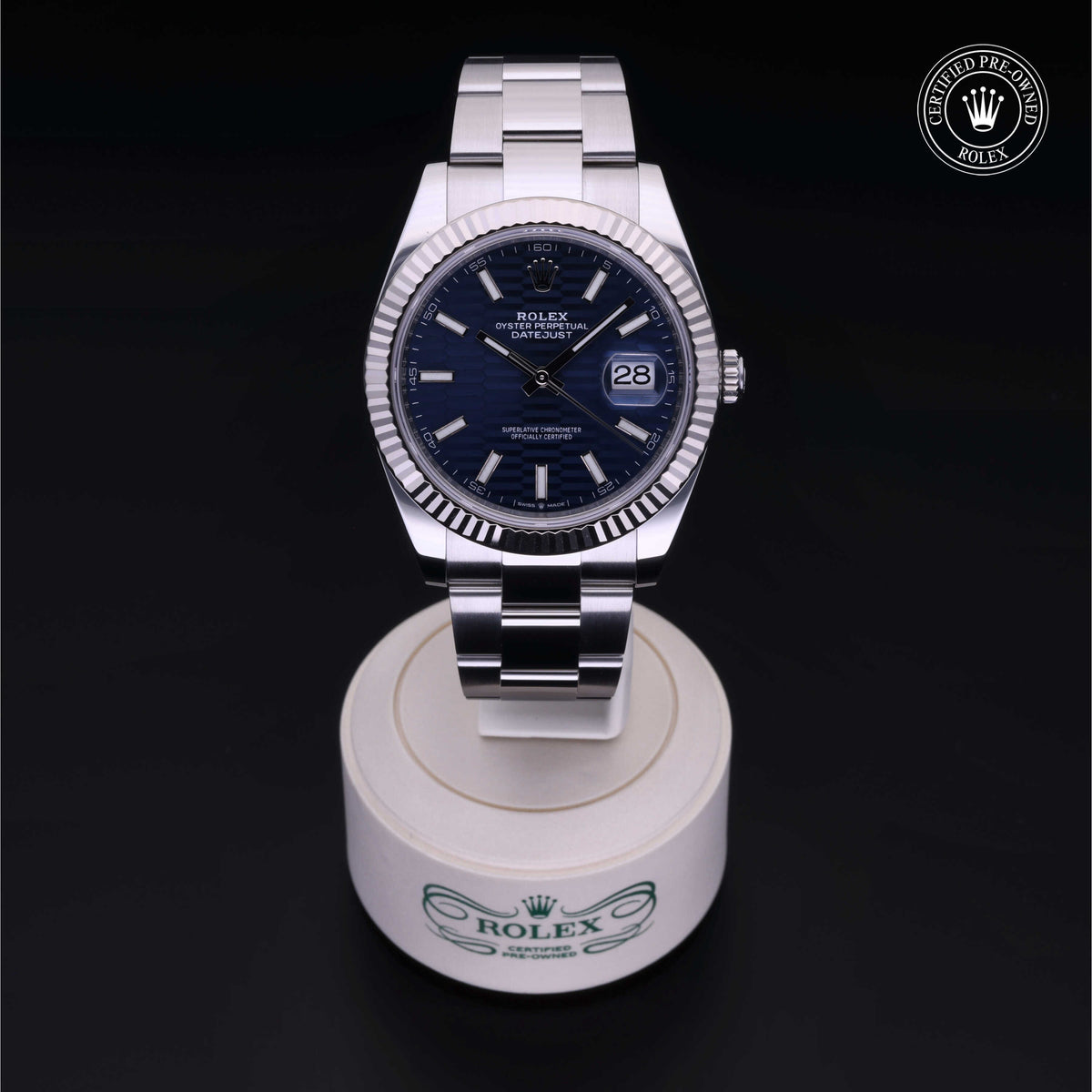 A Rolex Datejust watch with a silver metal bracelet and deep blue dial, displayed on a branded stand with a Rolex Certified Pre-Owned logo above, highlighting luxury watch craftsmanship.