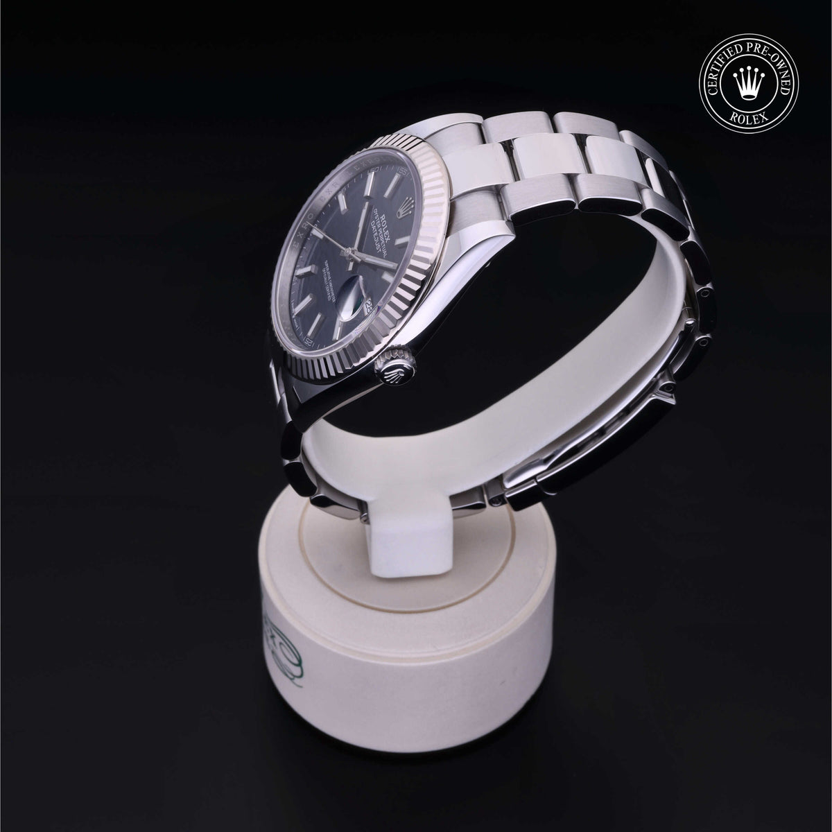 Silver Rolex wristwatch with a black dial and fluted bezel displayed on a round stand against a dark background, featuring a Certified Pre-Owned Rolex logo.