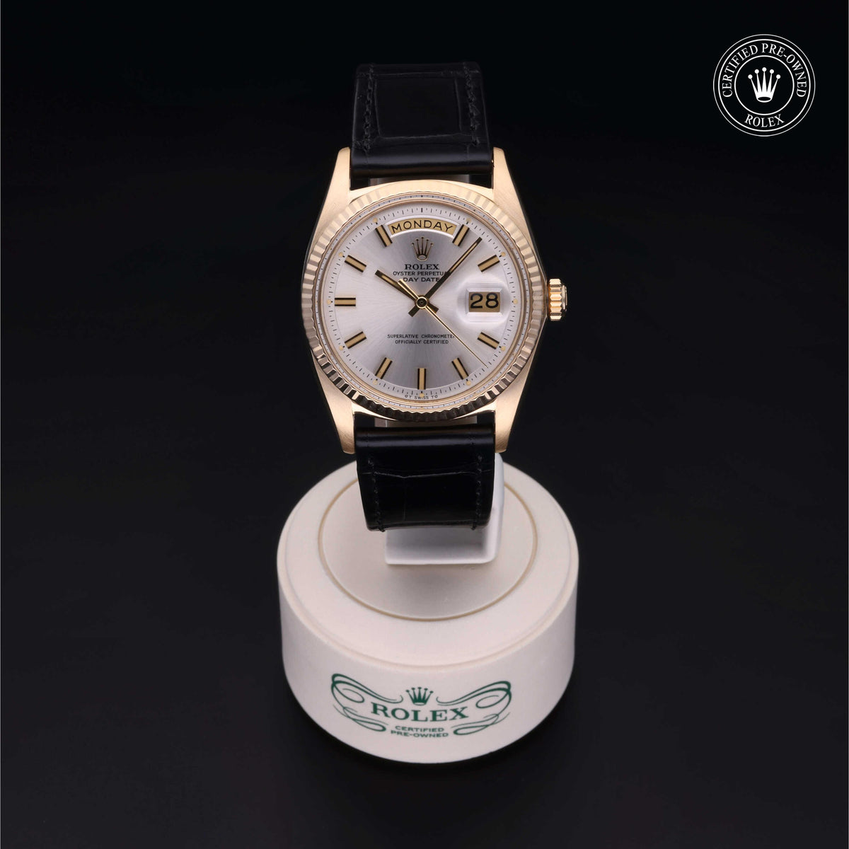 Luxury Rolex watch with gold bezel and black leather strap displayed on a white branded stand, showcasing the day and date feature under elegant lighting.