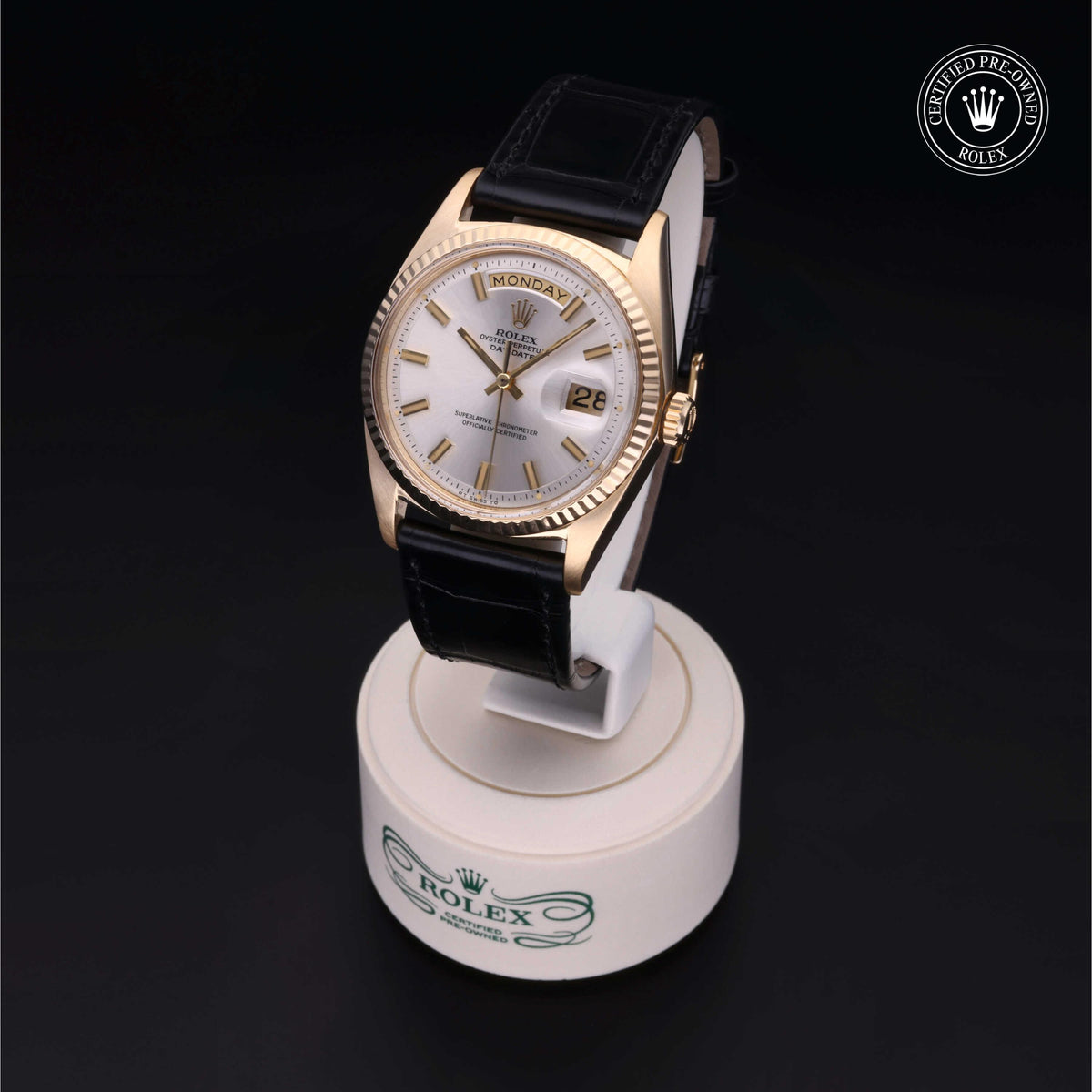 Rolex luxury watch with gold case, white dial, day and date display, black leather strap, displayed on a round stand with Rolex branding and certified pre-owned logo.