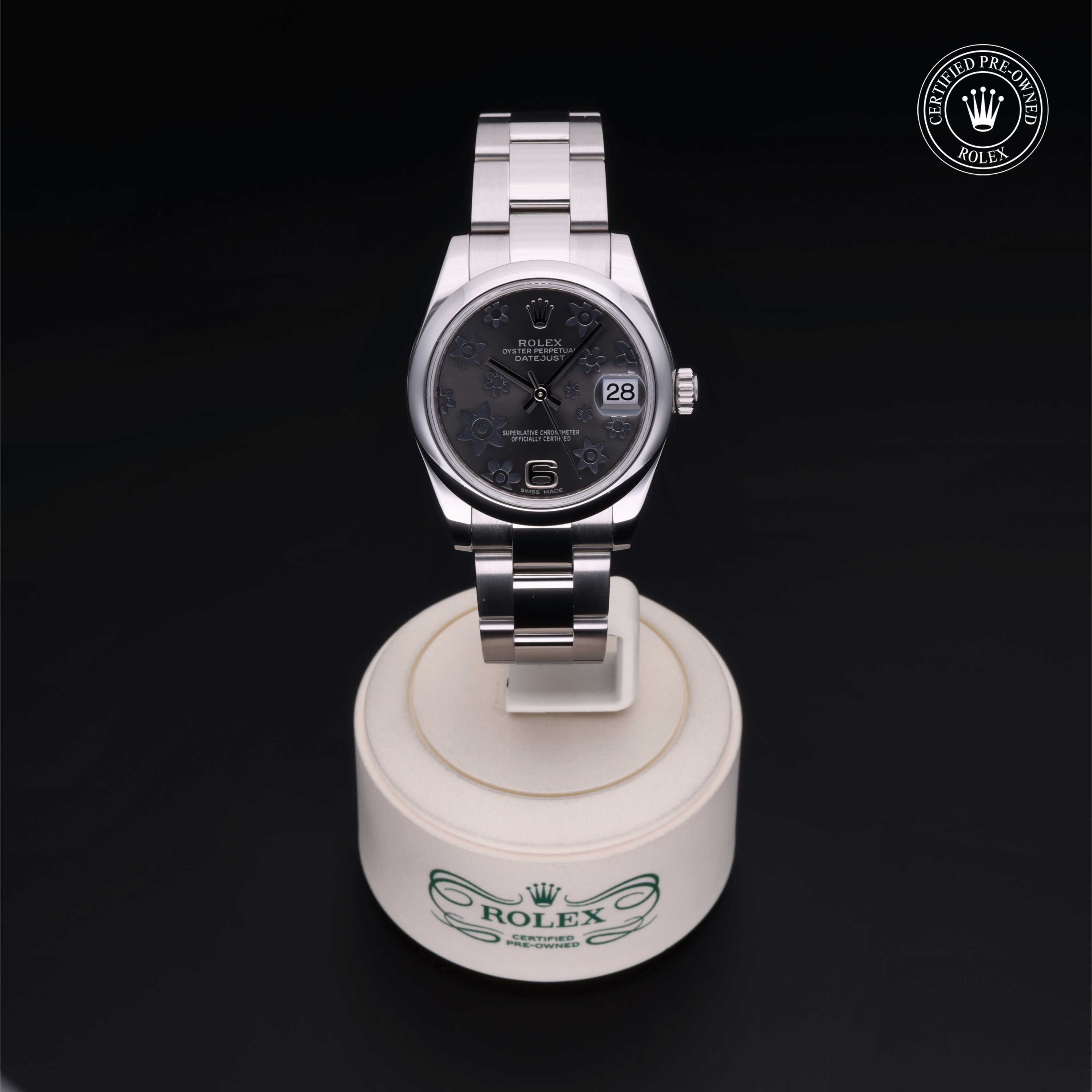 A silver Rolex luxury watch with a black patterned dial and date display on a white round stand, featuring the Rolex logo and a certified pre-owned emblem in the corner.