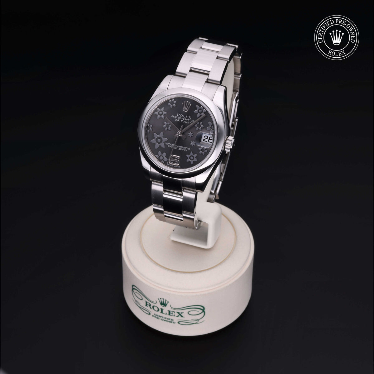 Stainless steel Rolex watch with a black floral-patterned dial and date display on a white circular stand with the Rolex logo, set against a black background.