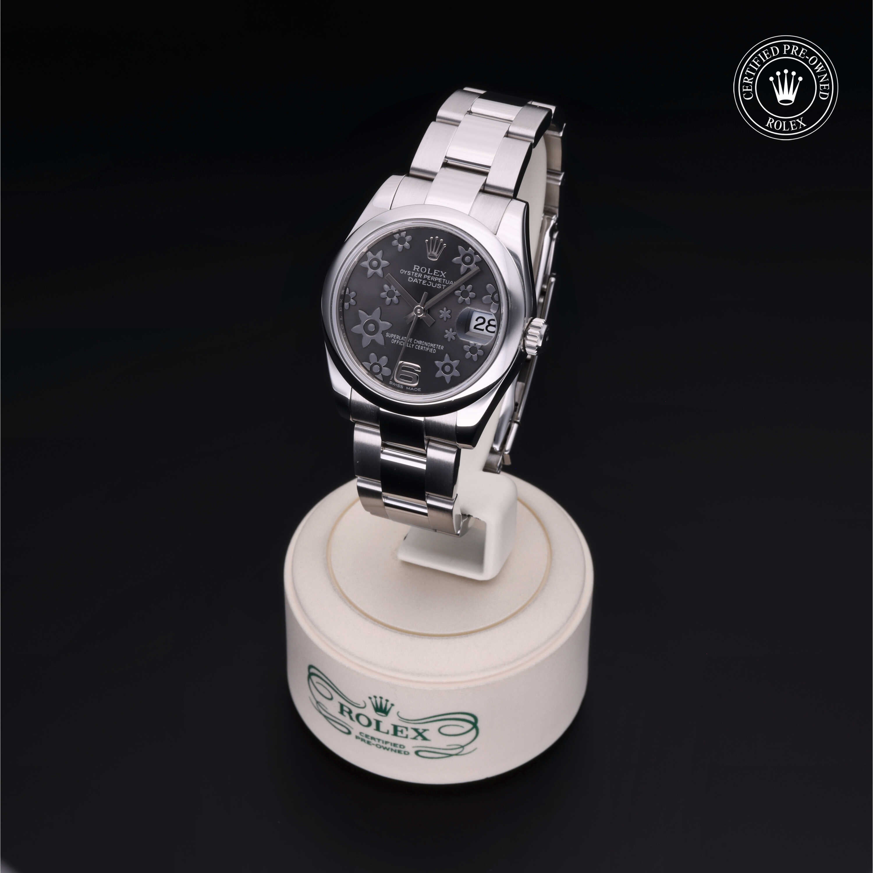 Stainless steel Rolex watch with a black floral-patterned dial and date display on a white circular stand with the Rolex logo, set against a black background.