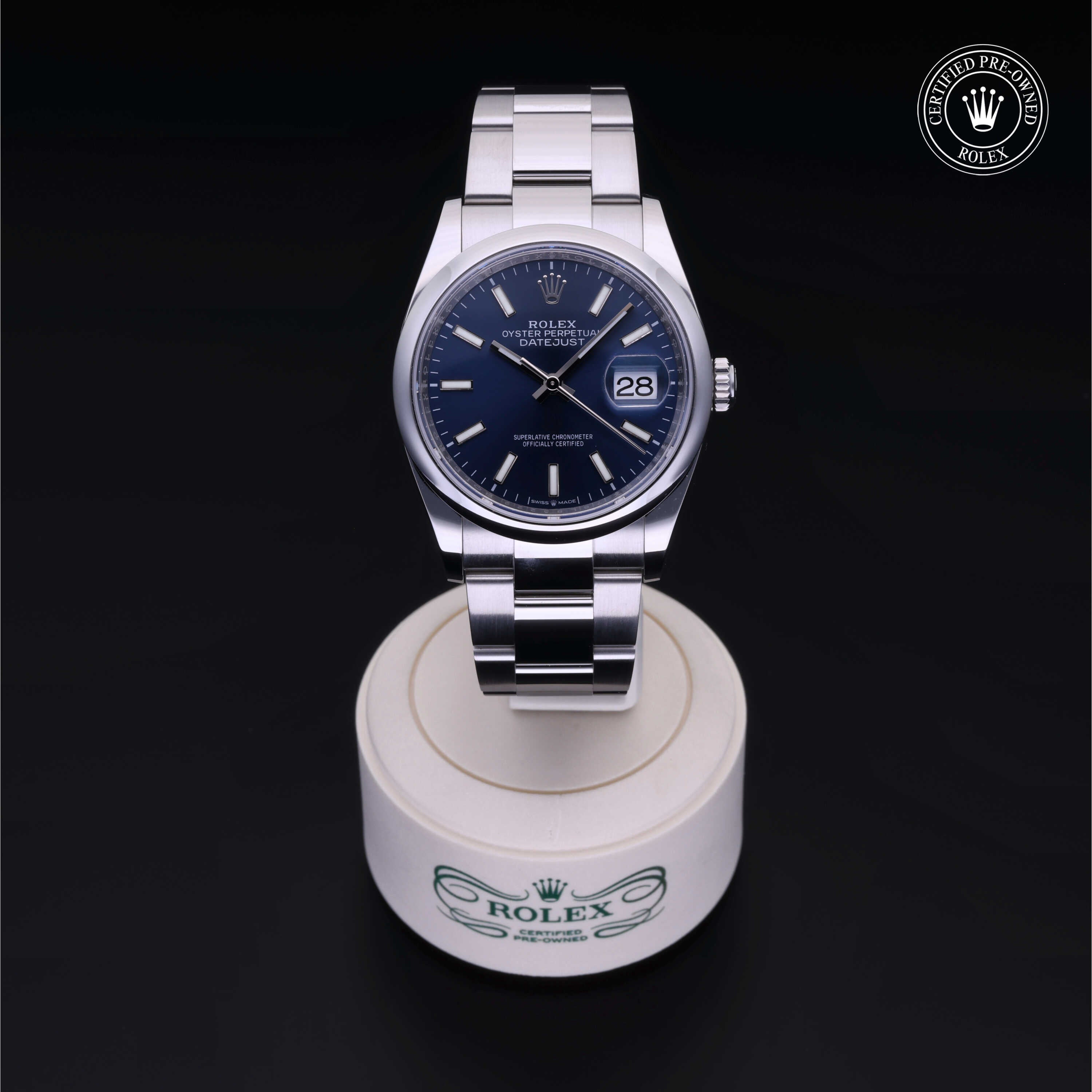 Rolex Datejust watch with a blue dial and silver stainless steel bracelet displayed on a white stand with Rolex branding, showing the date at 3 o'clock.