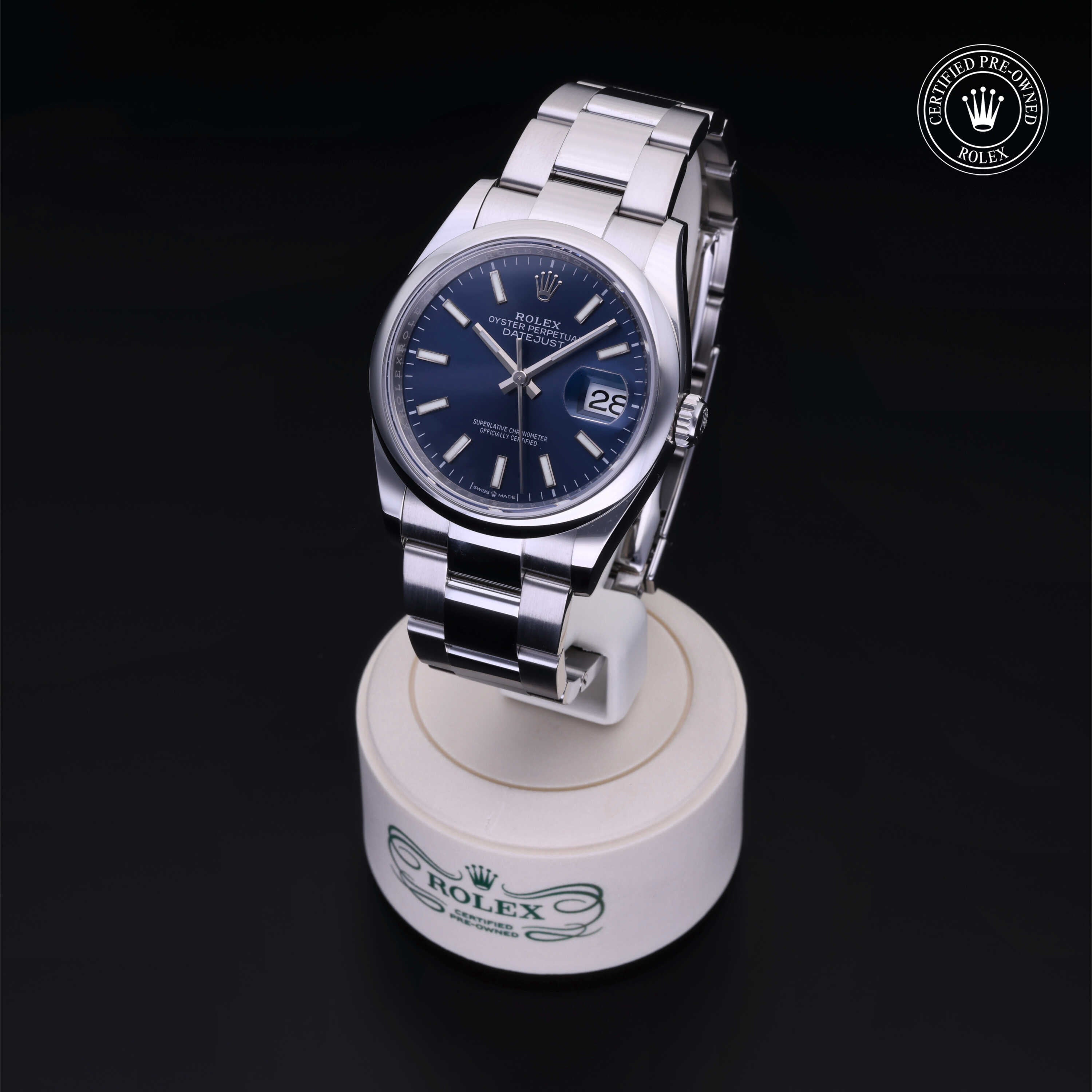 Rolex luxury watch with blue dial and stainless steel band displayed on a white stand, featuring date window and certified pre-owned Rolex logo in the corner.