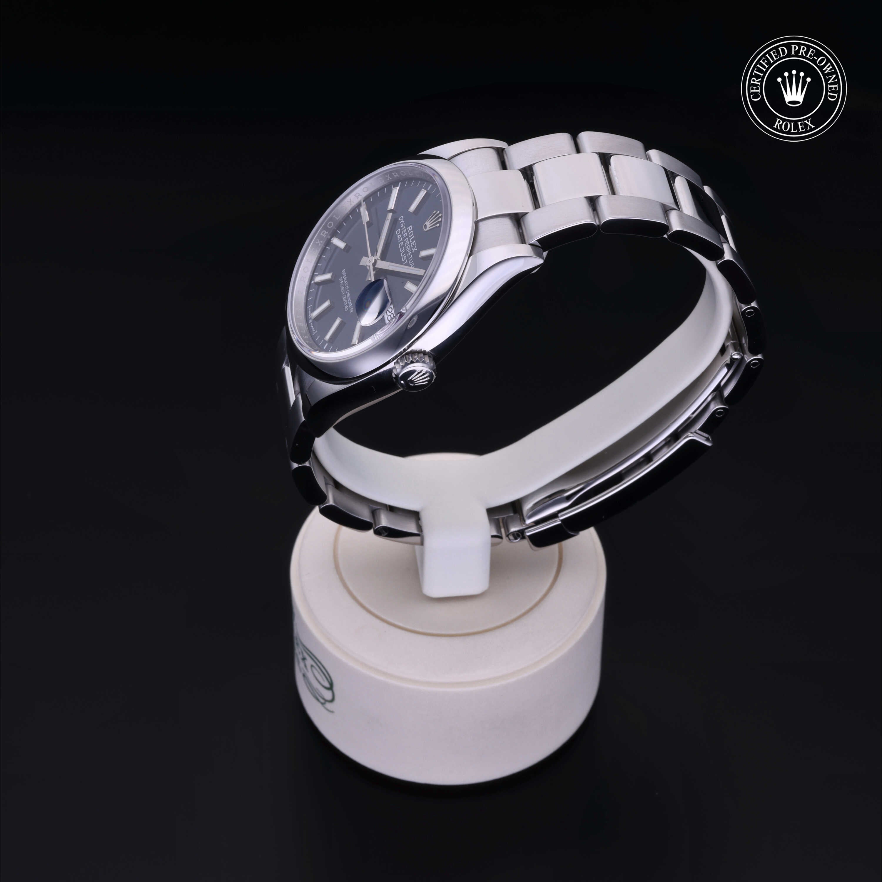 Side view of a luxury Rolex Certified Pre-Owned stainless steel watch with a dark dial, displayed on a white stand against a black background.