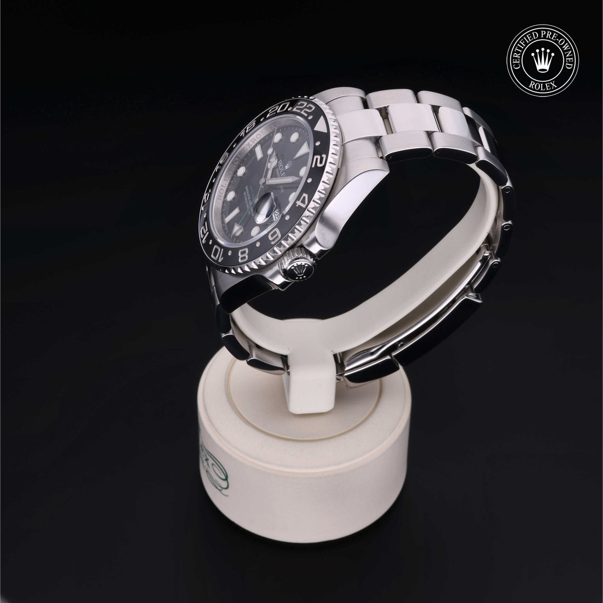 A stainless steel Rolex watch with a black dial and bezel, displayed on a white circular stand against a black background, featuring a certified pre-owned Rolex logo.