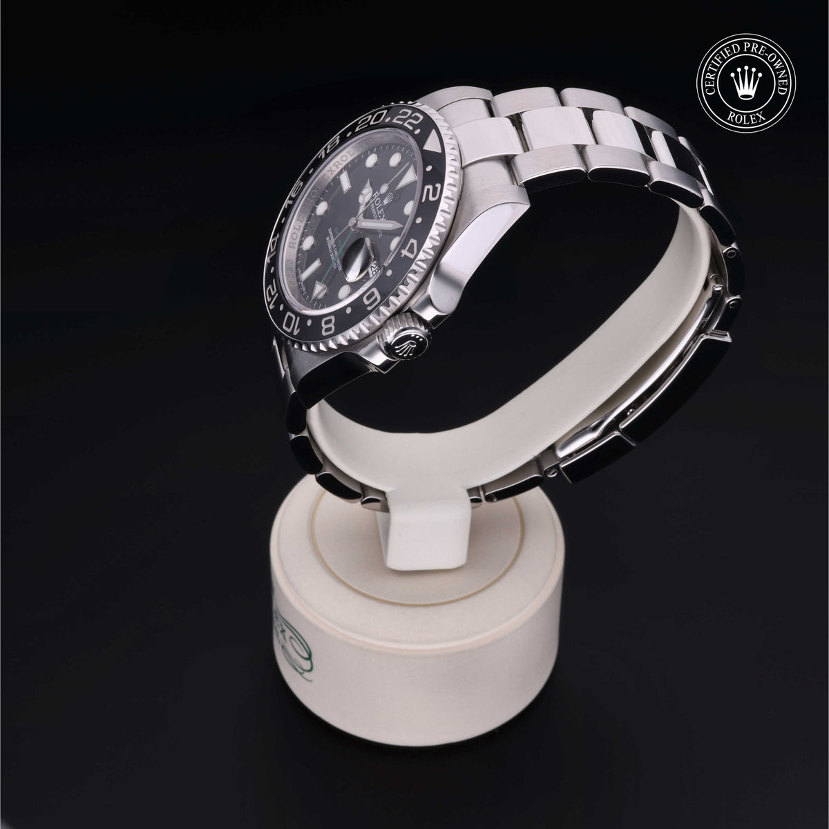 A stainless steel luxury watch with a black dial and rotating bezel displayed on a white stand against a black background, featuring a certified pre-owned Rolex logo.