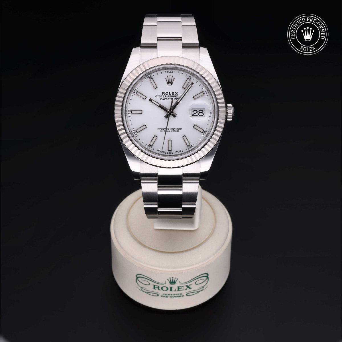 A luxury Rolex Datejust watch with a silver metal band and white dial displayed on a branded circular stand, featuring a date window and certified pre-owned Rolex logo nearby.
