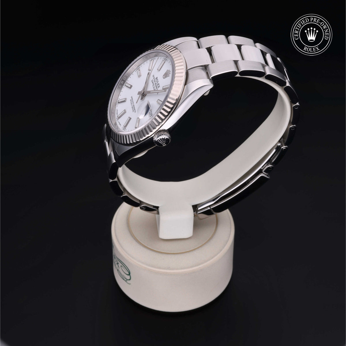 A silver Rolex luxury watch with a white dial and fluted bezel displayed on a white round stand against a black background, featuring a certified pre-owned Rolex emblem.