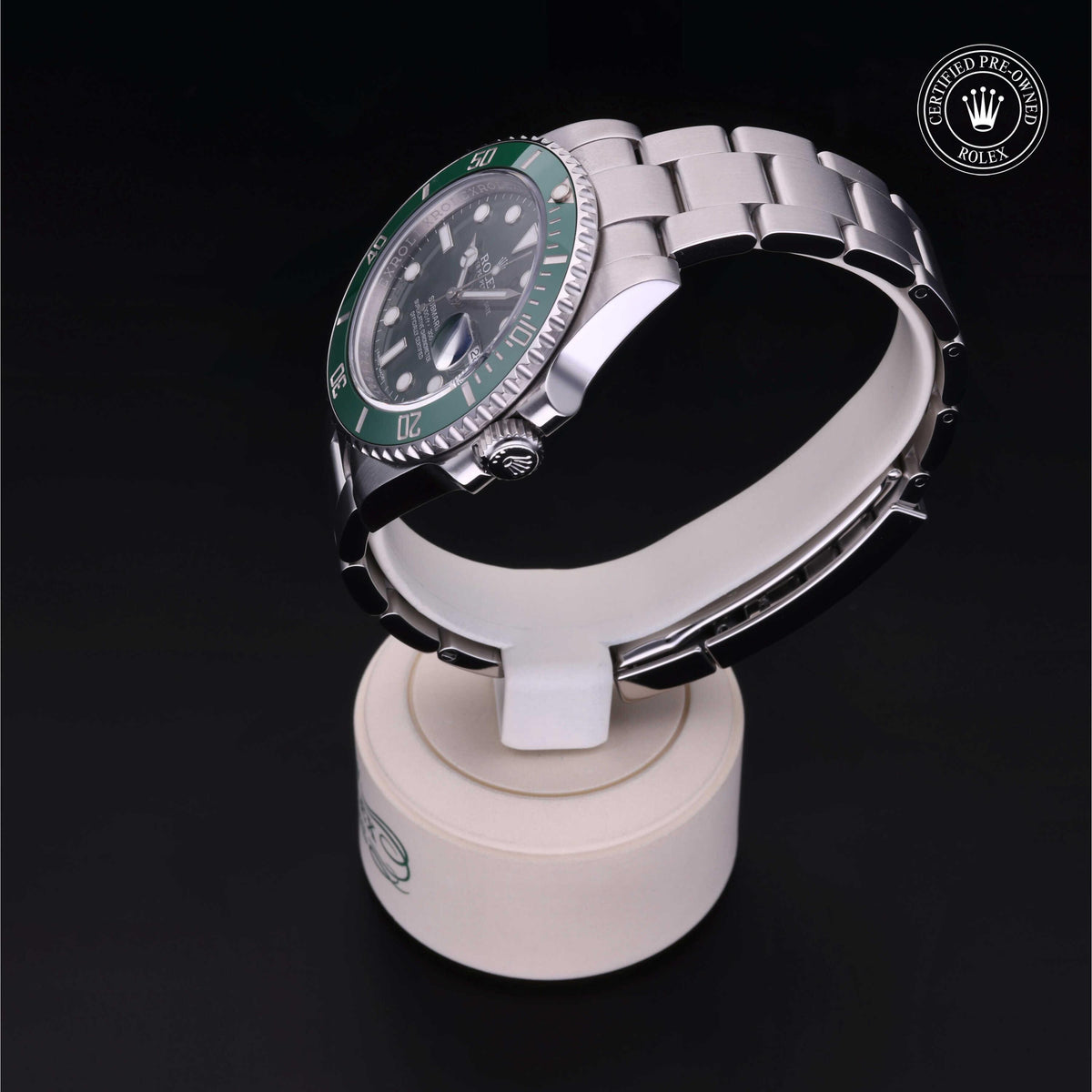 Silver luxury watch with a green bezel displayed on a white stand against a black background, featuring a certified pre-owned Rolex emblem.