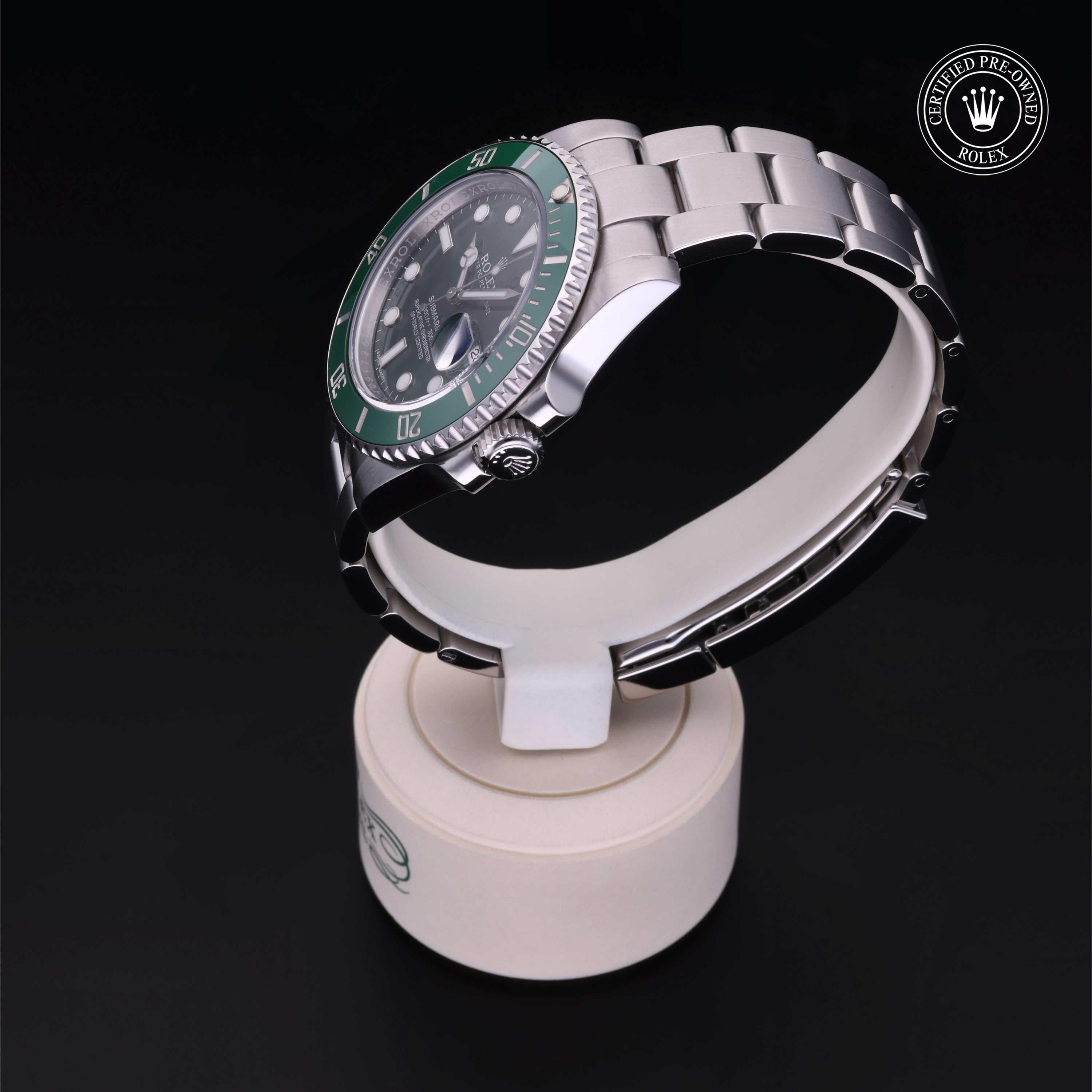 Silver luxury watch with a green bezel displayed on a white stand against a black background, featuring a certified pre-owned Rolex emblem.