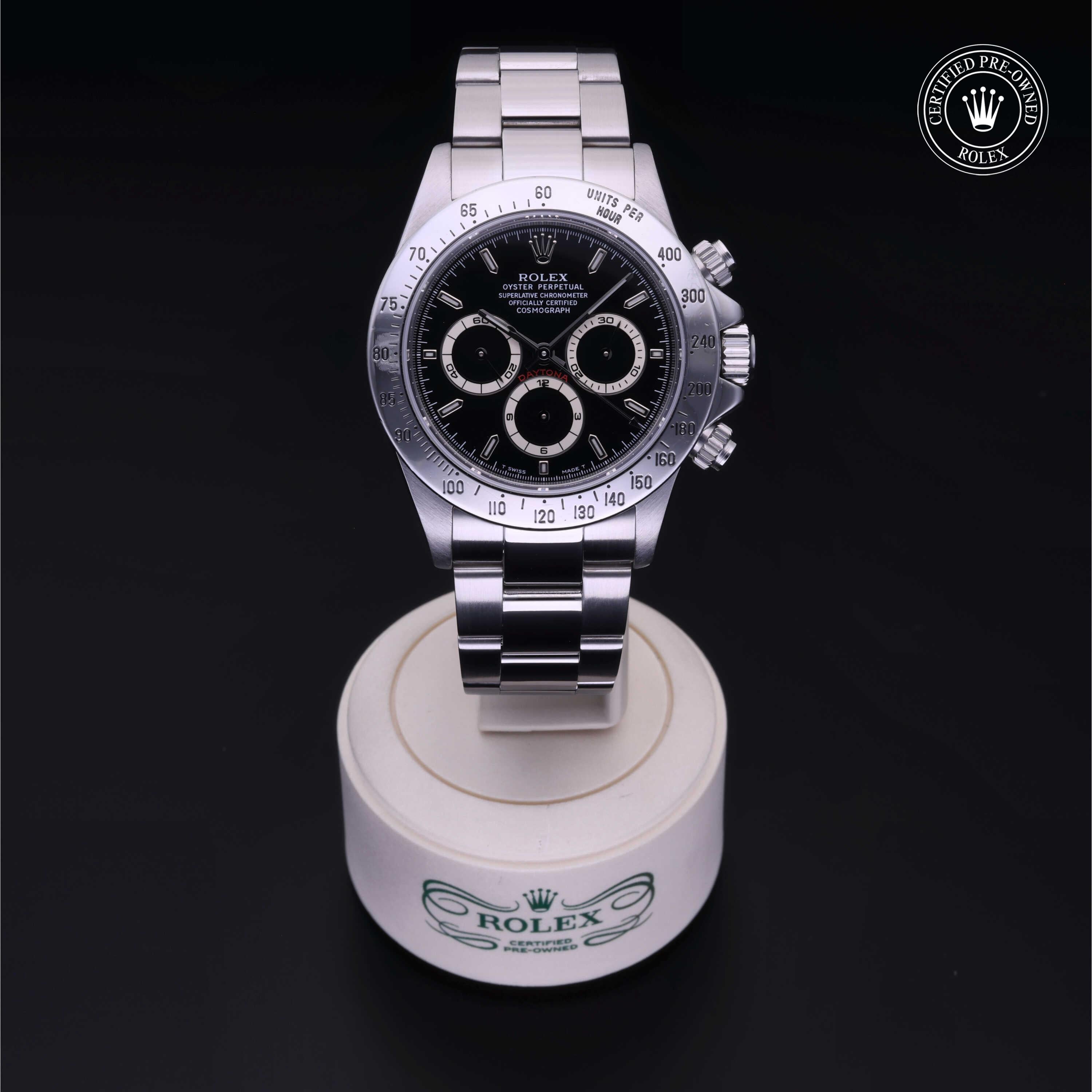 A stainless steel Rolex chronograph watch with a black dial and three subdials displayed on a white stand, featuring the Rolex logo and a certified pre-owned emblem.