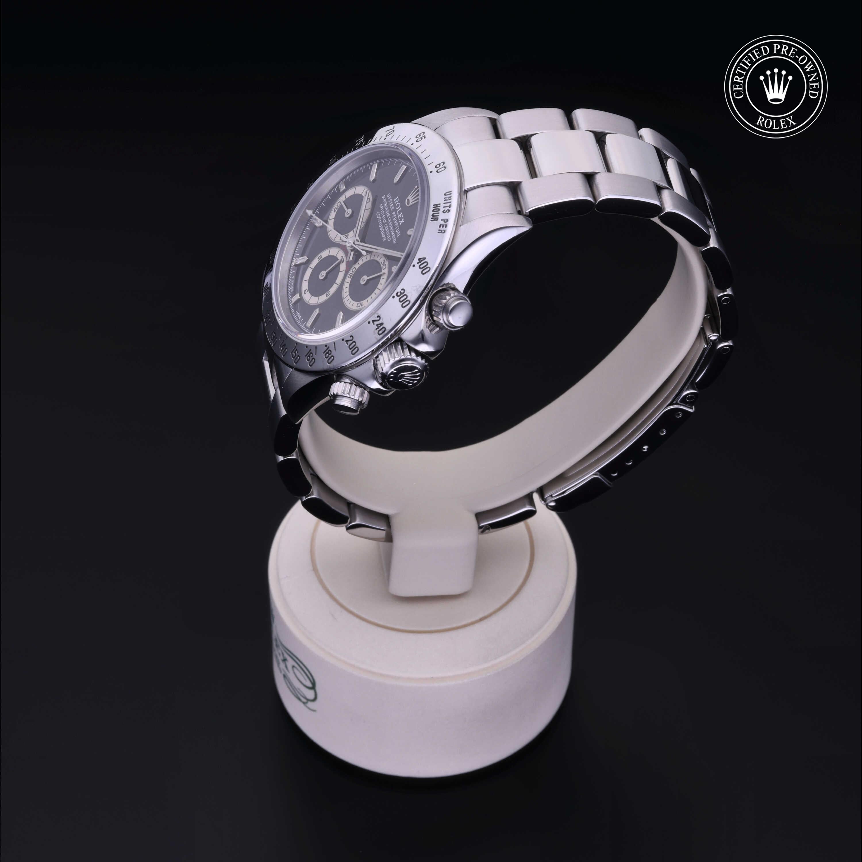 A luxury stainless steel Rolex chronograph watch with a black dial displayed on a white stand against a black background, featuring the Rolex Certified Pre-Owned emblem.