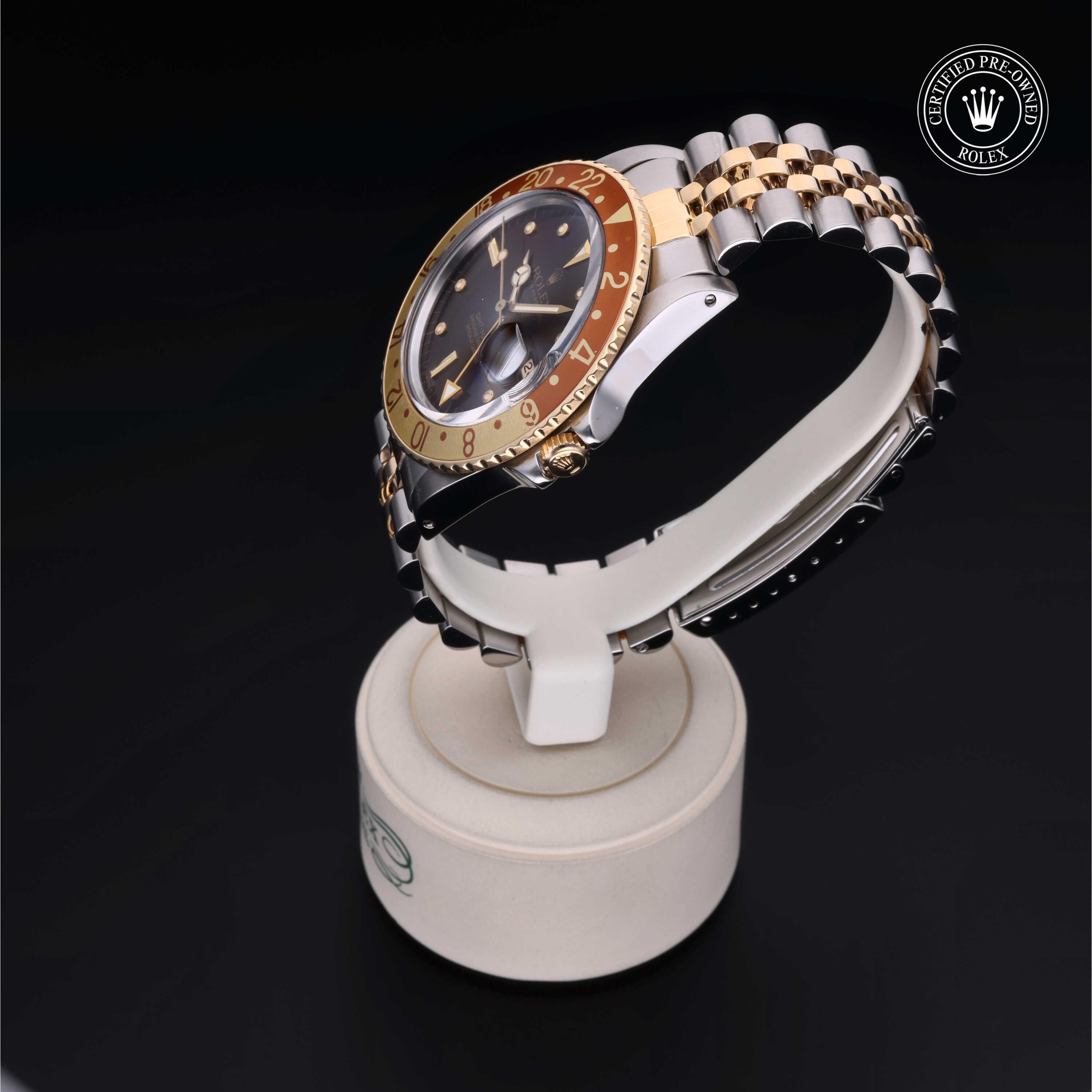 Two-tone Rolex wristwatch with a gold and silver bracelet displayed on a white stand, featuring a dark dial and a rotating bezel with gold and brown tones, certified pre-owned.