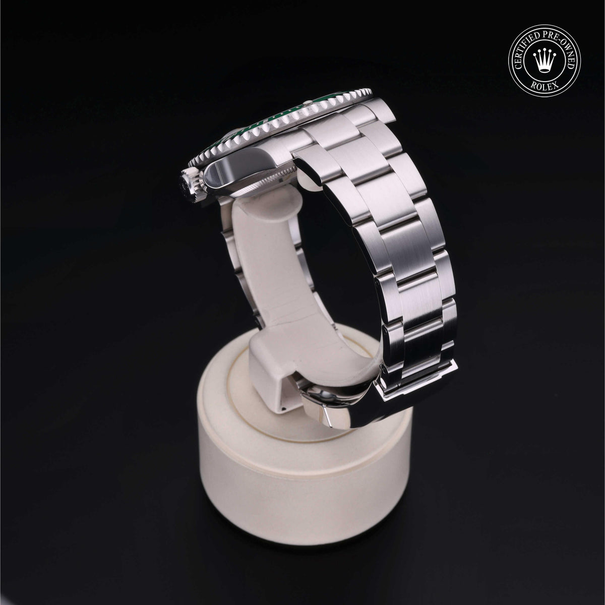 Close-up of a luxury stainless steel Rolex watch with a polished bracelet, displayed on a white stand against a black background, featuring a certified pre-owned Rolex logo.