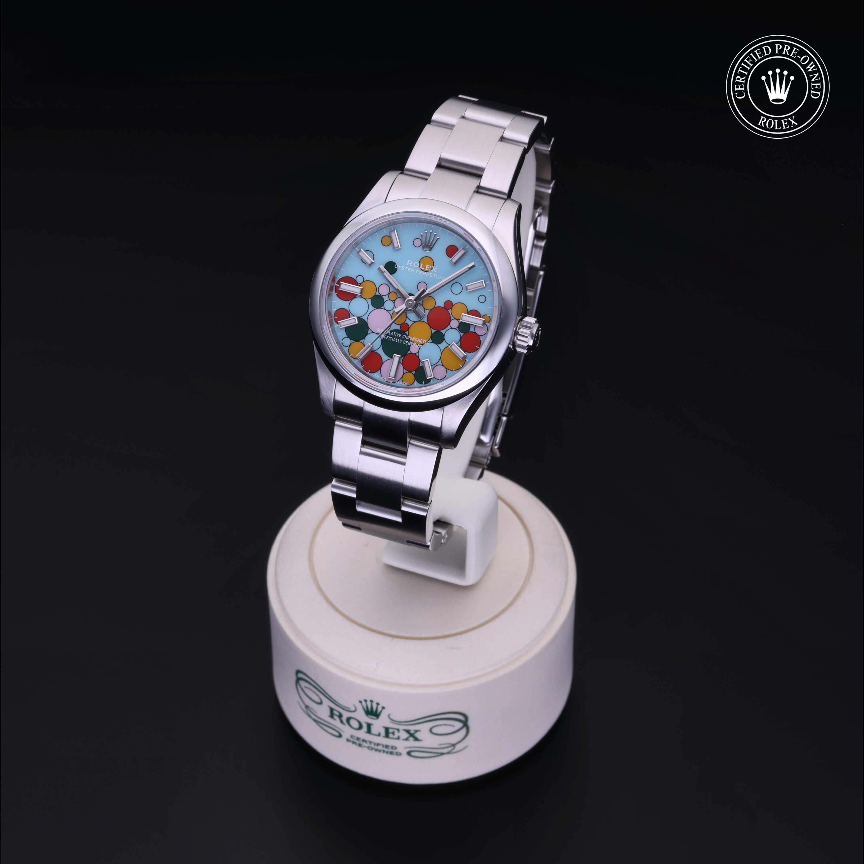 Rolex luxury watch with a stainless steel band and a colorful dial featuring vibrant circular patterns, displayed on a branded stand with a certified pre-owned Rolex logo.