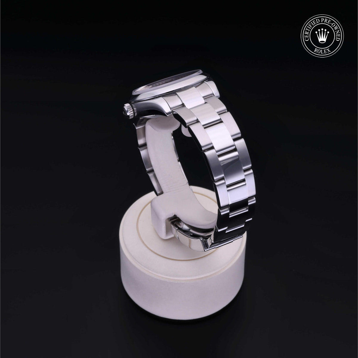 Side view of a polished stainless steel Rolex luxury watch displayed on a white cushion stand with a certified pre-owned Rolex logo in the corner, showcasing fine craftsmanship.