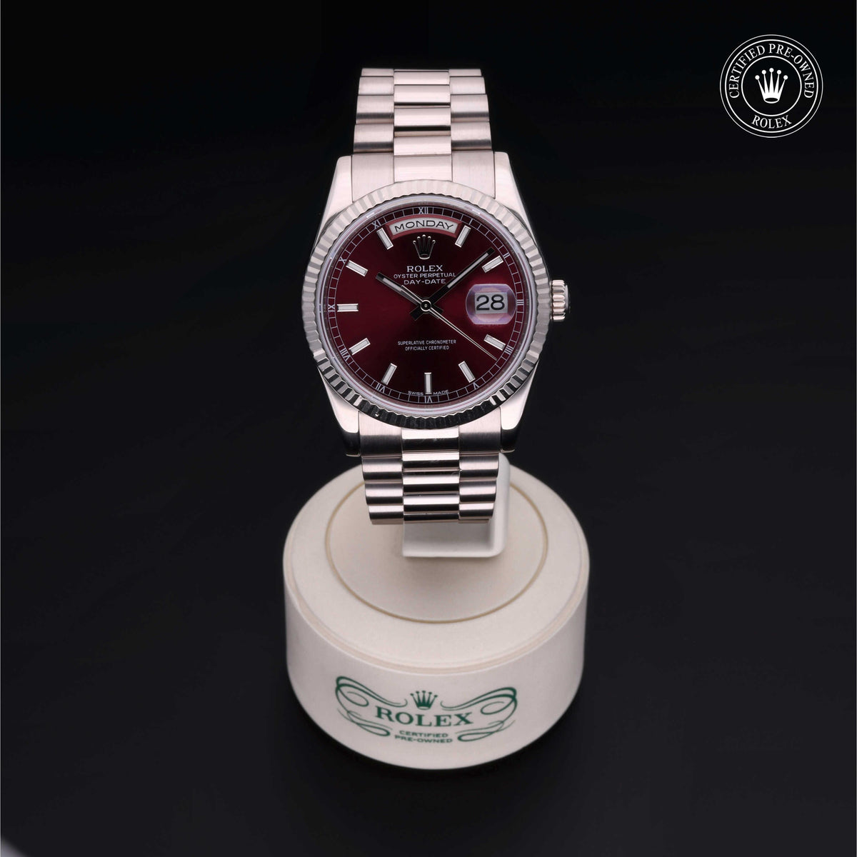 Close-up of a Rolex wristwatch with a burgundy dial and silver bracelet, displayed on a white circular stand with the Rolex logo, featuring a certified pre-owned badge.