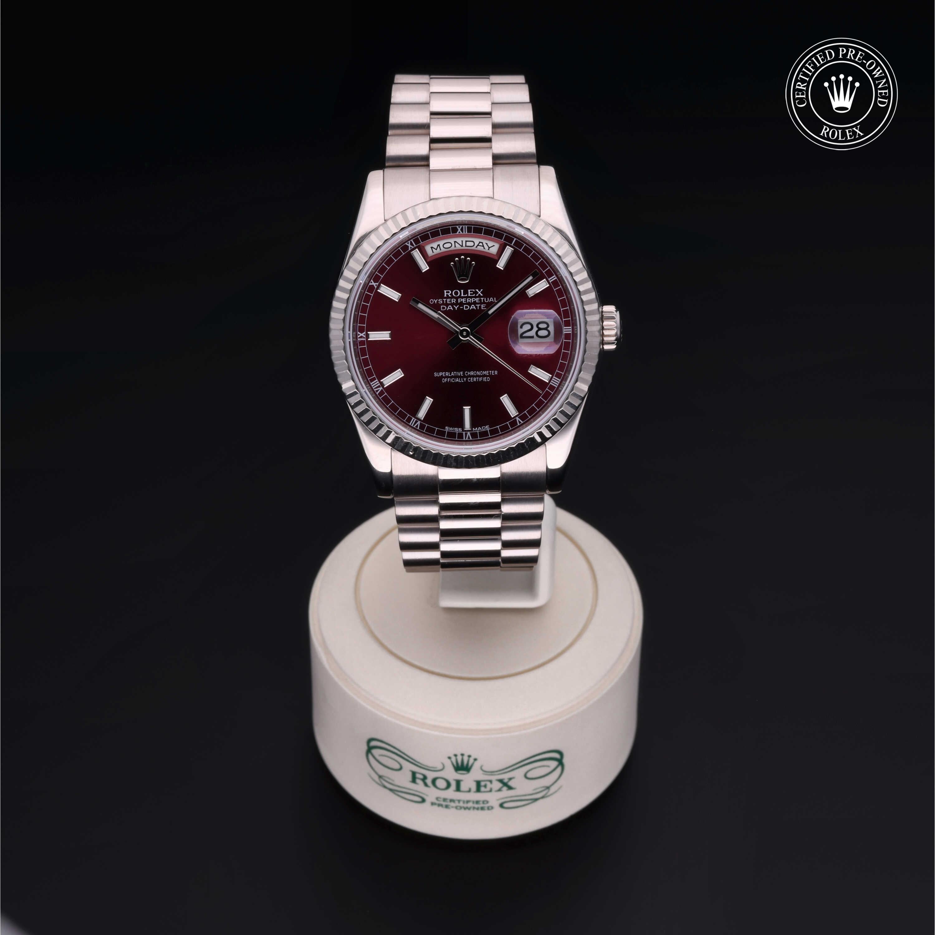 Close-up of a Rolex wristwatch with a burgundy dial and silver bracelet, displayed on a white circular stand with the Rolex logo, featuring a certified pre-owned badge.