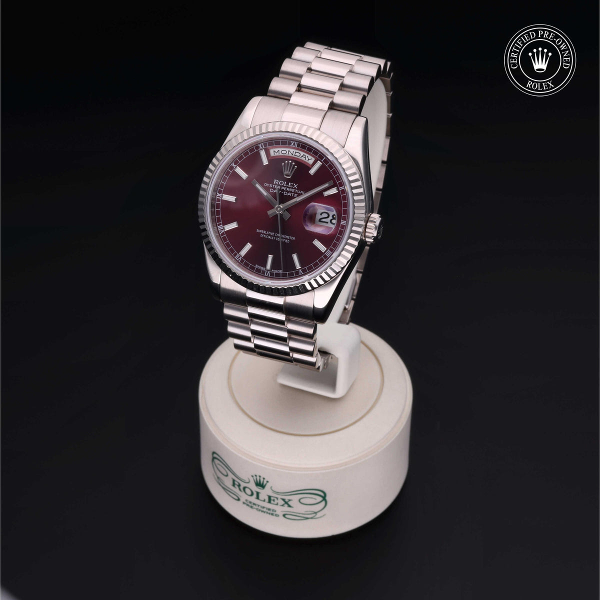Rolex luxury watch with a burgundy dial and silver bracelet displayed on a white stand against a dark background, featuring a certified pre-owned Rolex emblem.