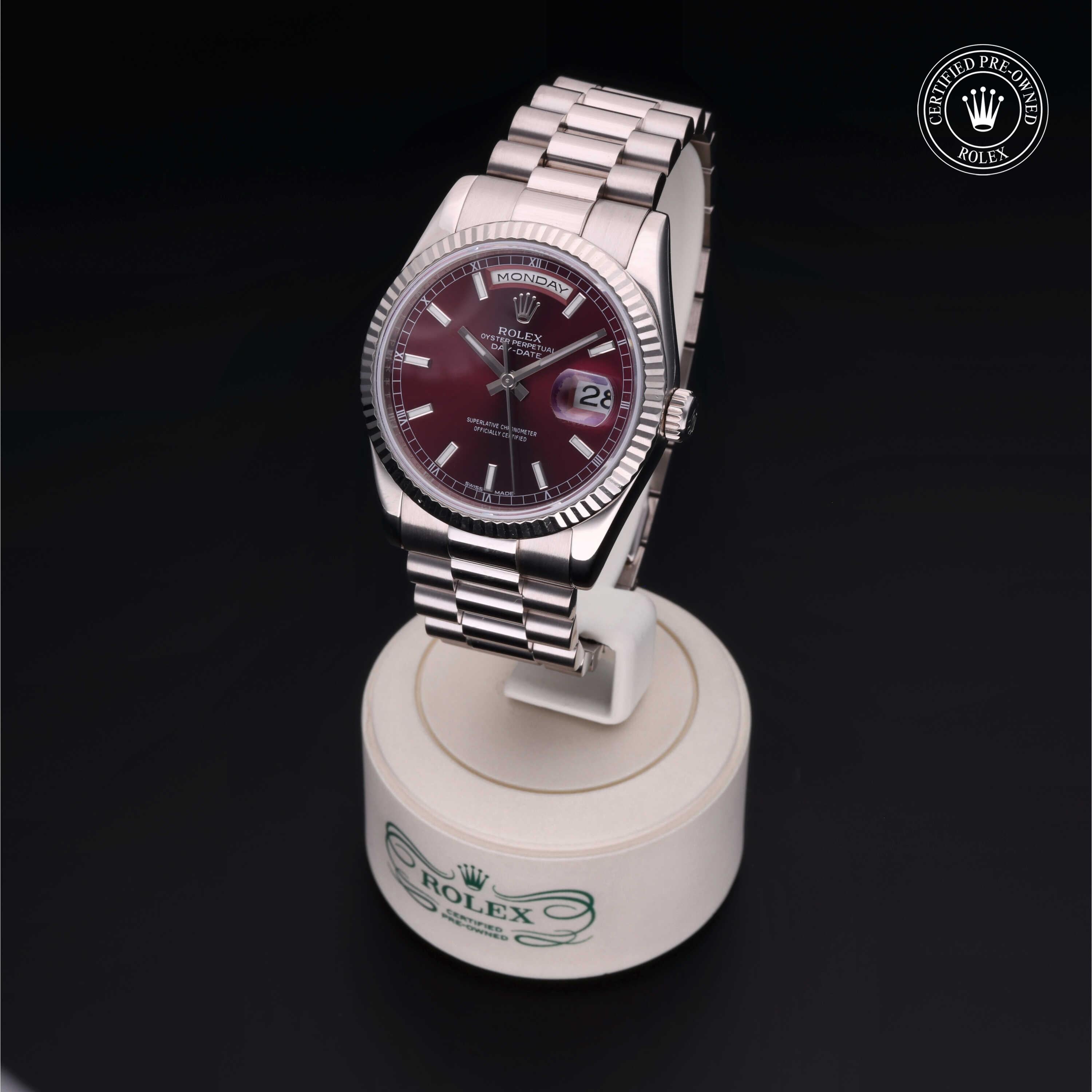 Rolex luxury watch with a burgundy dial and silver bracelet displayed on a white stand against a dark background, featuring a certified pre-owned Rolex emblem.