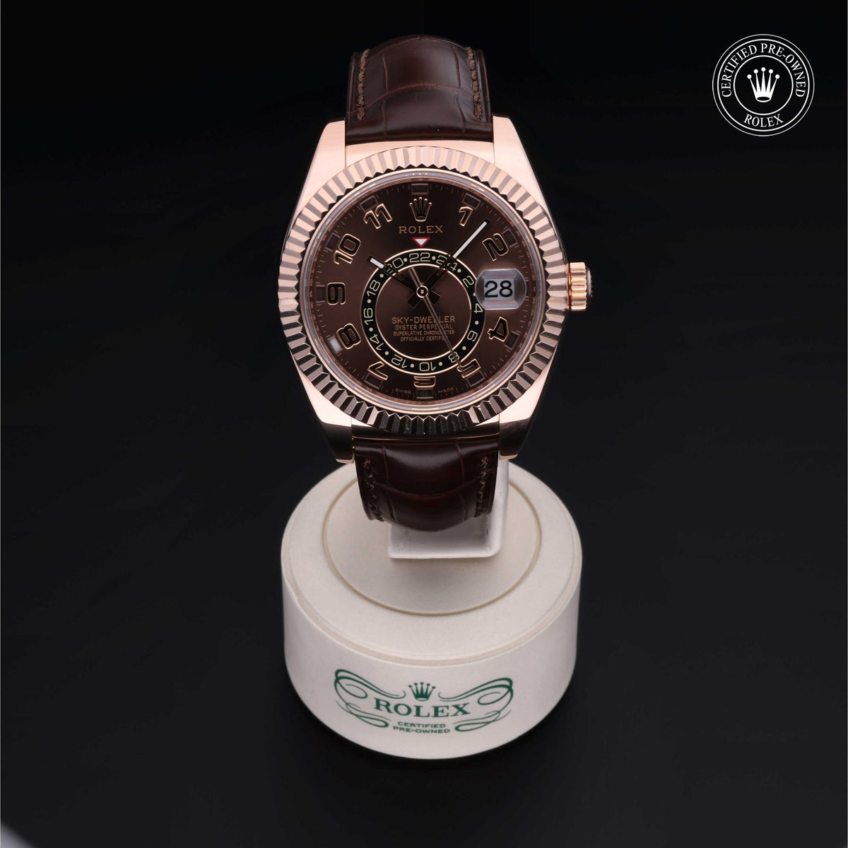 Rose gold Rolex watch with a brown leather strap displayed on a white stand with the Rolex logo, featuring a dark dial with date and time indicators.