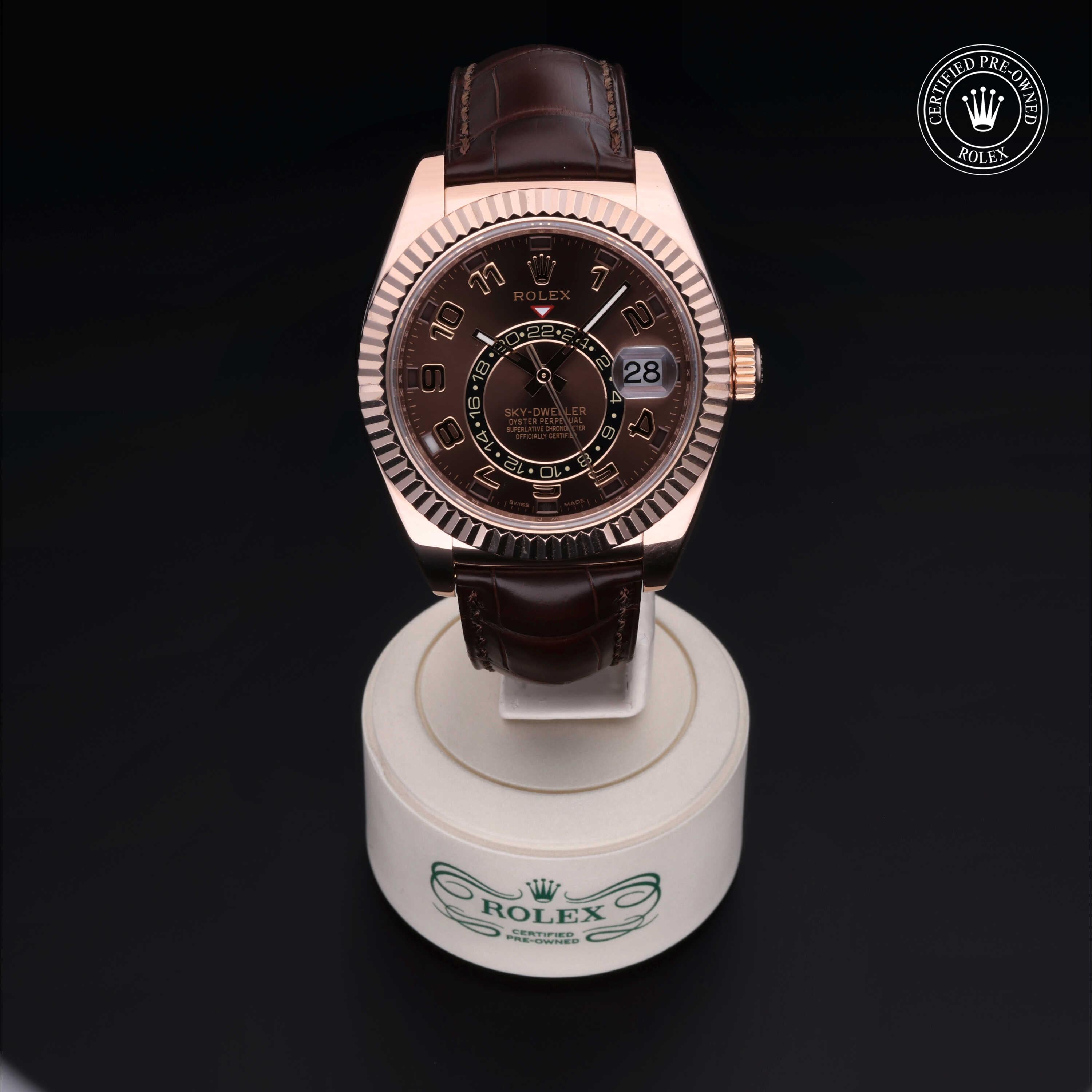 Rose gold Rolex watch with a brown leather strap displayed on a white stand with the Rolex logo, featuring a dark dial with date and time indicators.
