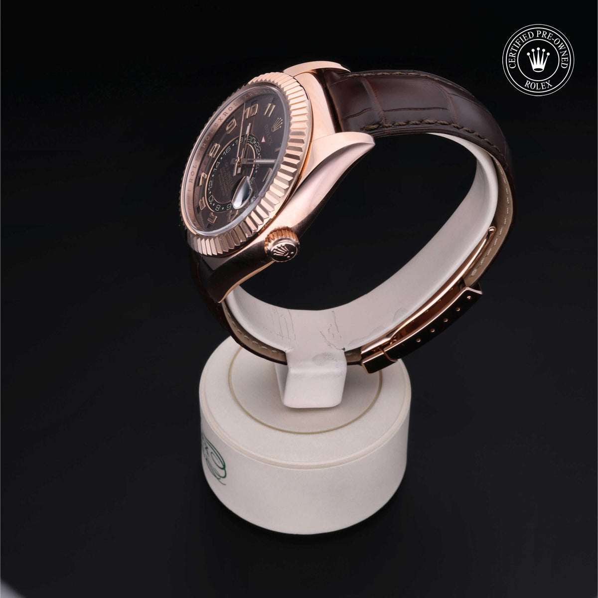 Side view of a luxury watch with a rose gold bezel and brown leather strap displayed on a white stand against a black background, featuring a certified pre-owned Rolex logo.