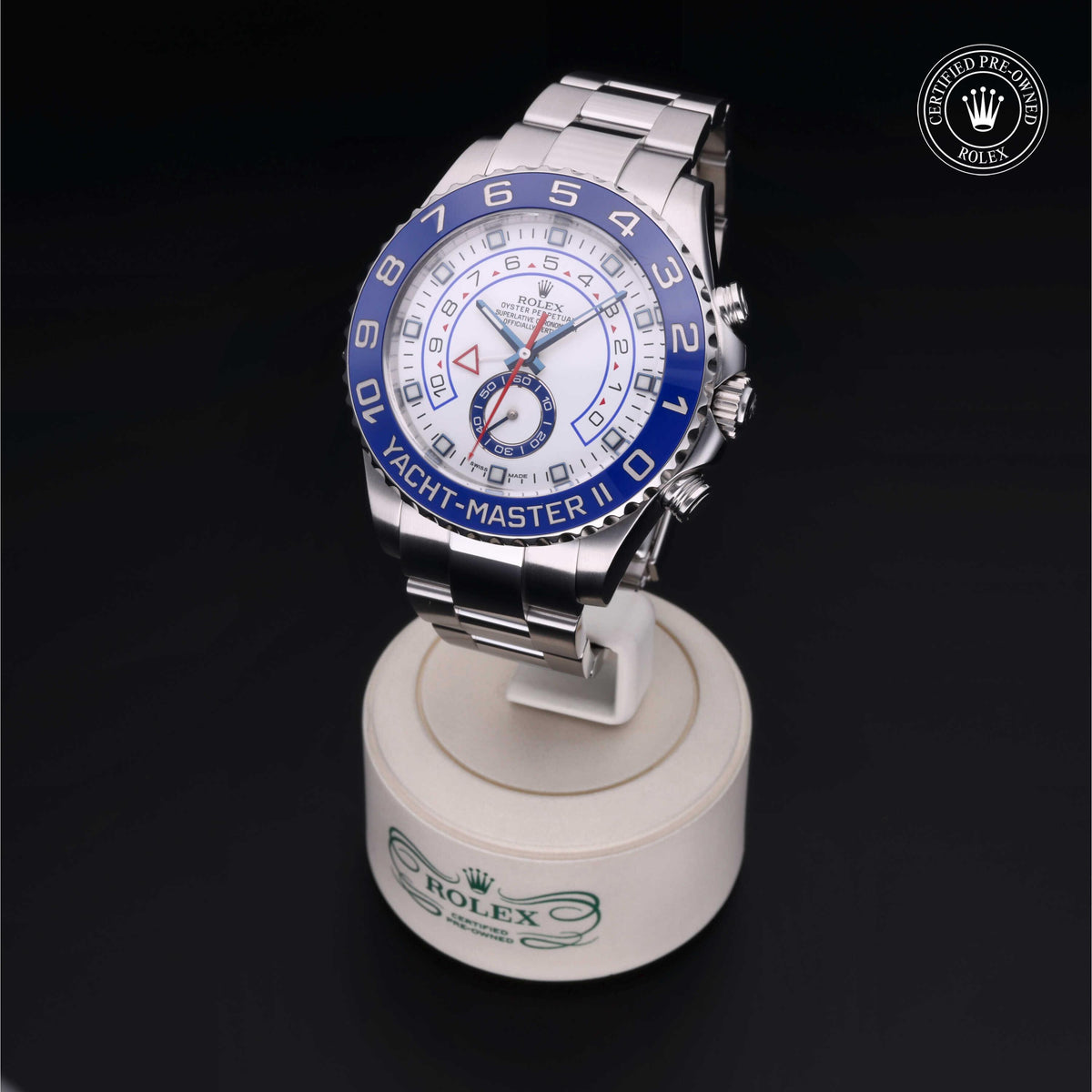 Silver Rolex Yacht-Master II watch with blue bezel and white dial displayed on a branded stand against a dark background, accompanied by a certified pre-owned Rolex logo.