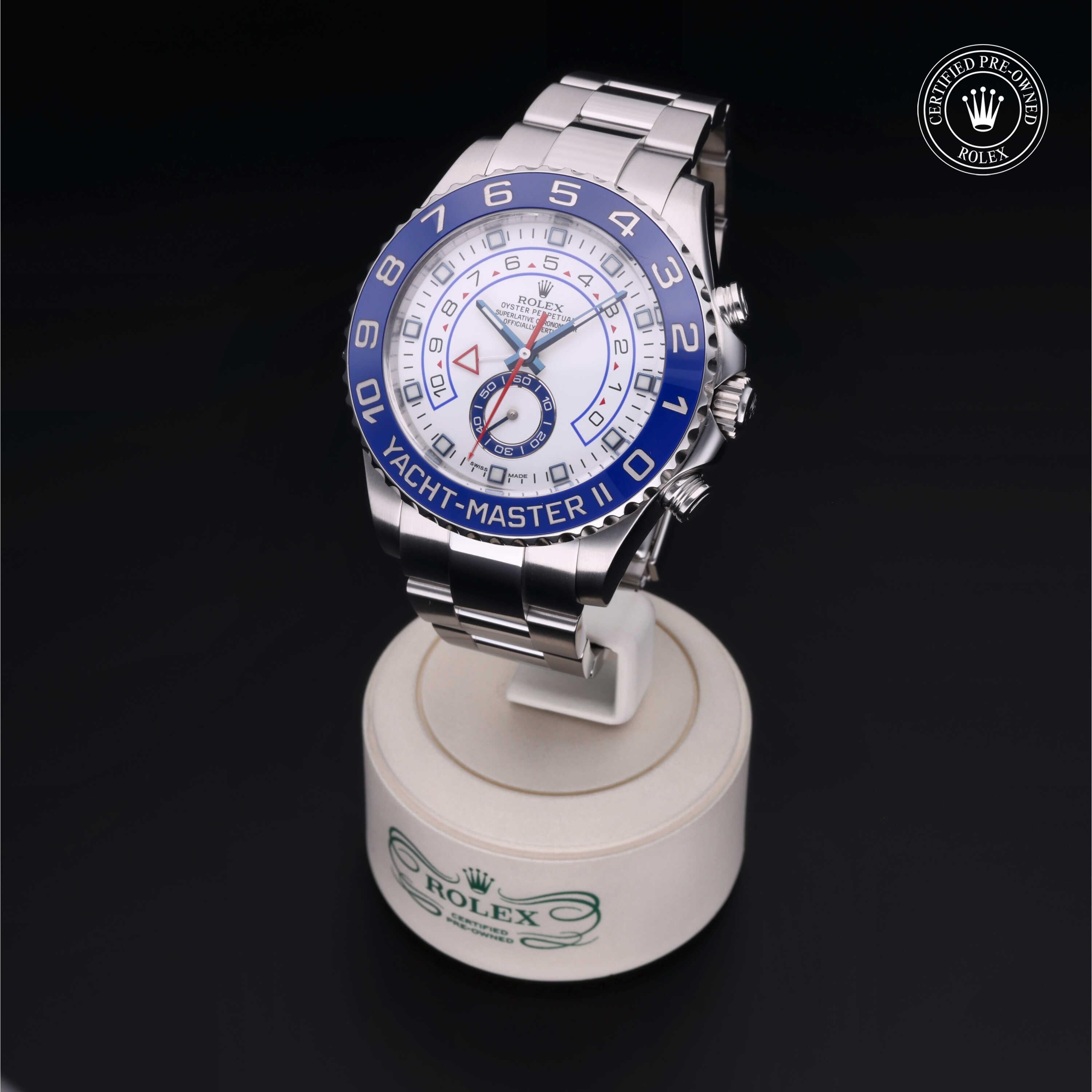 Silver Rolex Yacht-Master II watch with blue bezel and white dial displayed on a branded stand against a dark background, accompanied by a certified pre-owned Rolex logo.
