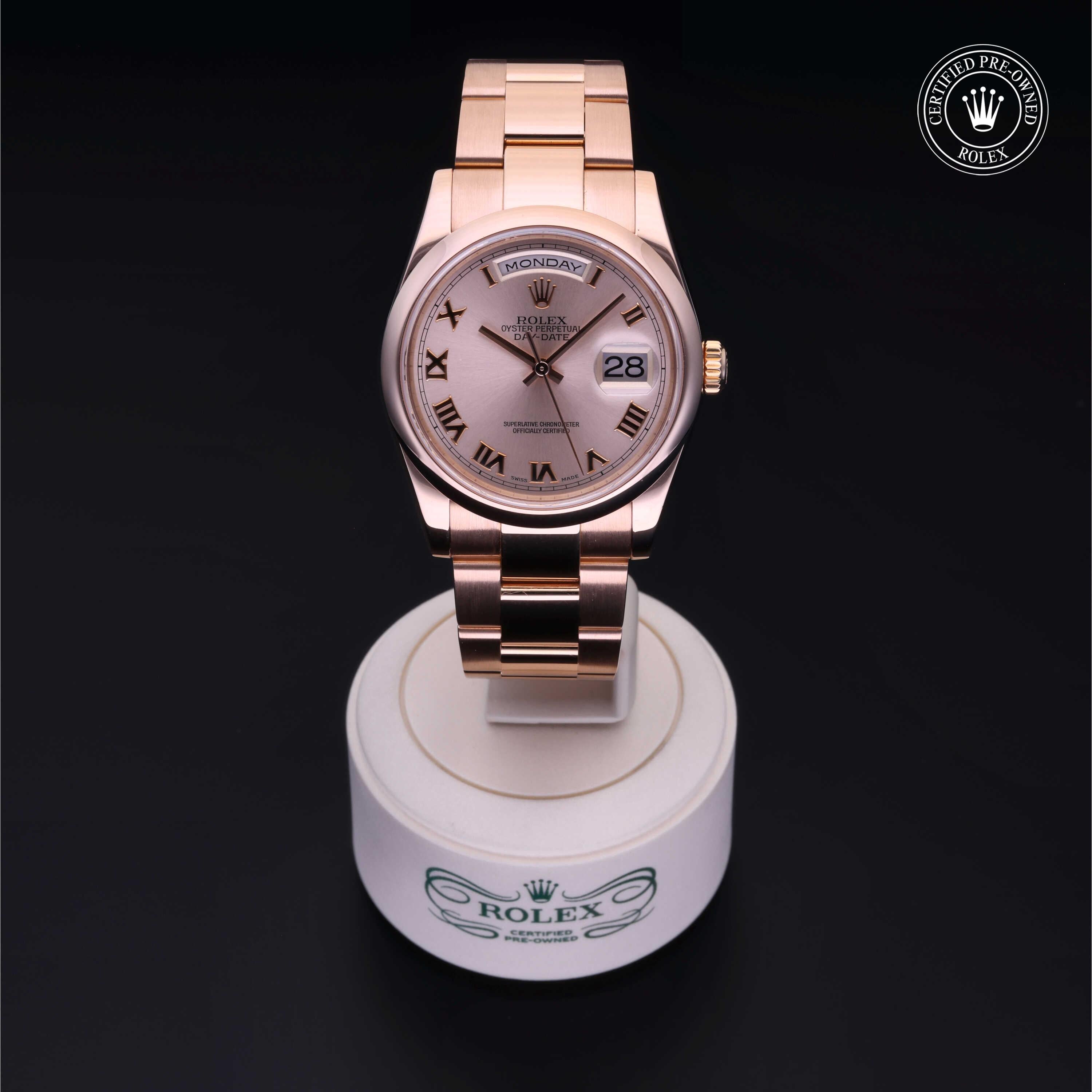 Rose gold Rolex watch with Roman numeral markers, day and date display, on a stand featuring the Rolex logo and Certified Pre-Owned emblem in the corner.