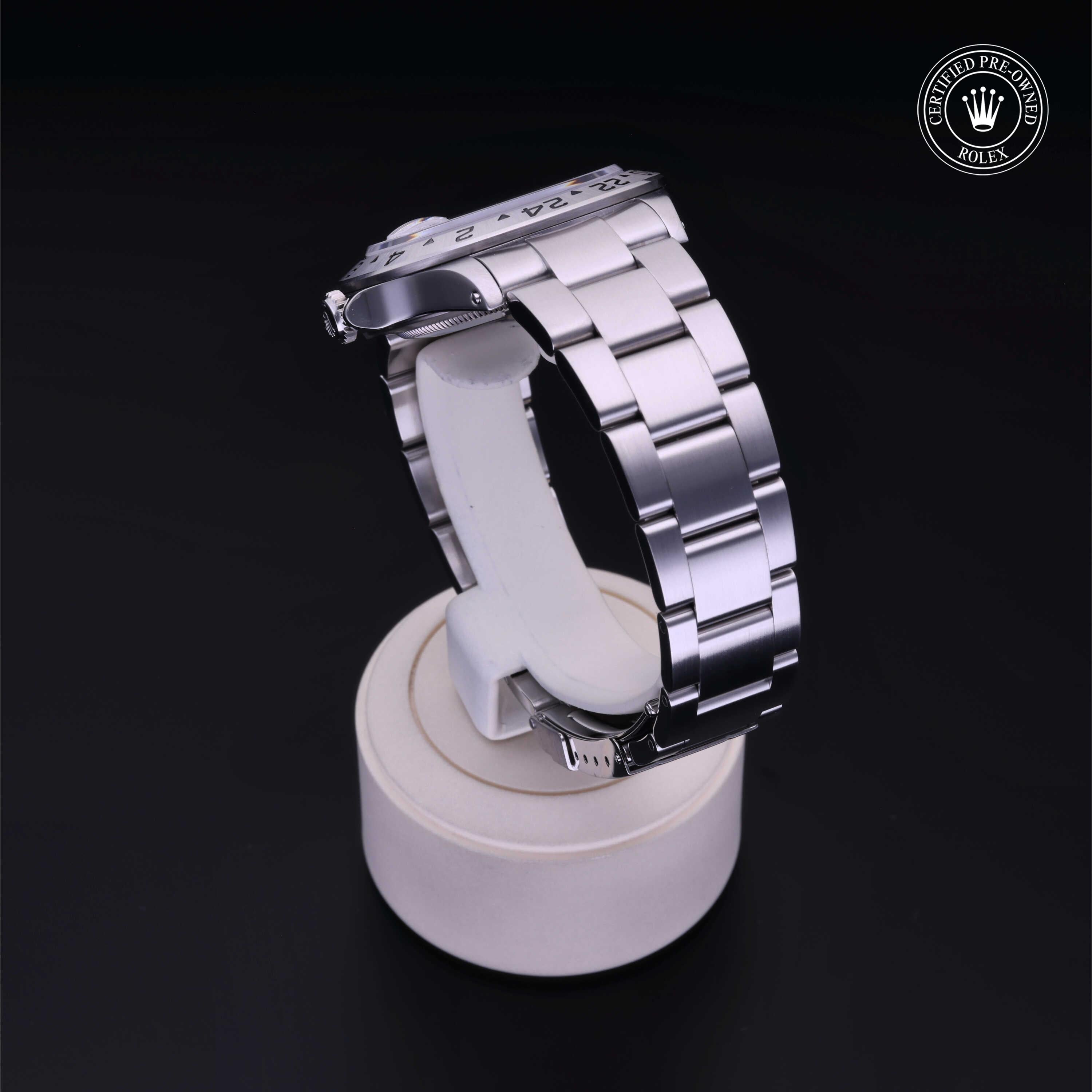 Close-up of a silver luxury watch with a polished metal band displayed on a white cylindrical stand against a black background, featuring a Rolex Certified Pre-Owned emblem.