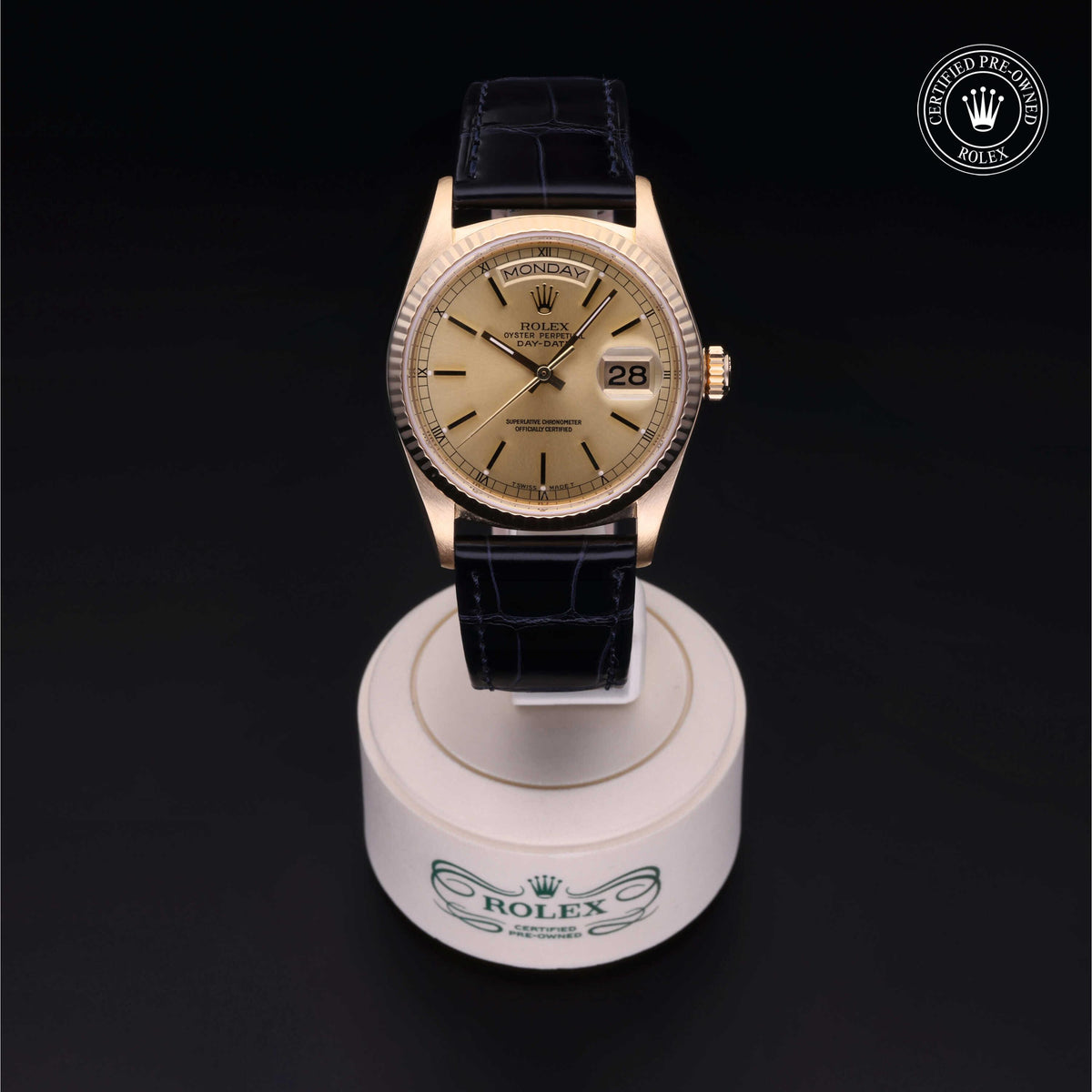 Luxurious gold Rolex watch with a beige dial, black leather strap, and day-date display, presented on a branded stand against a dark background.