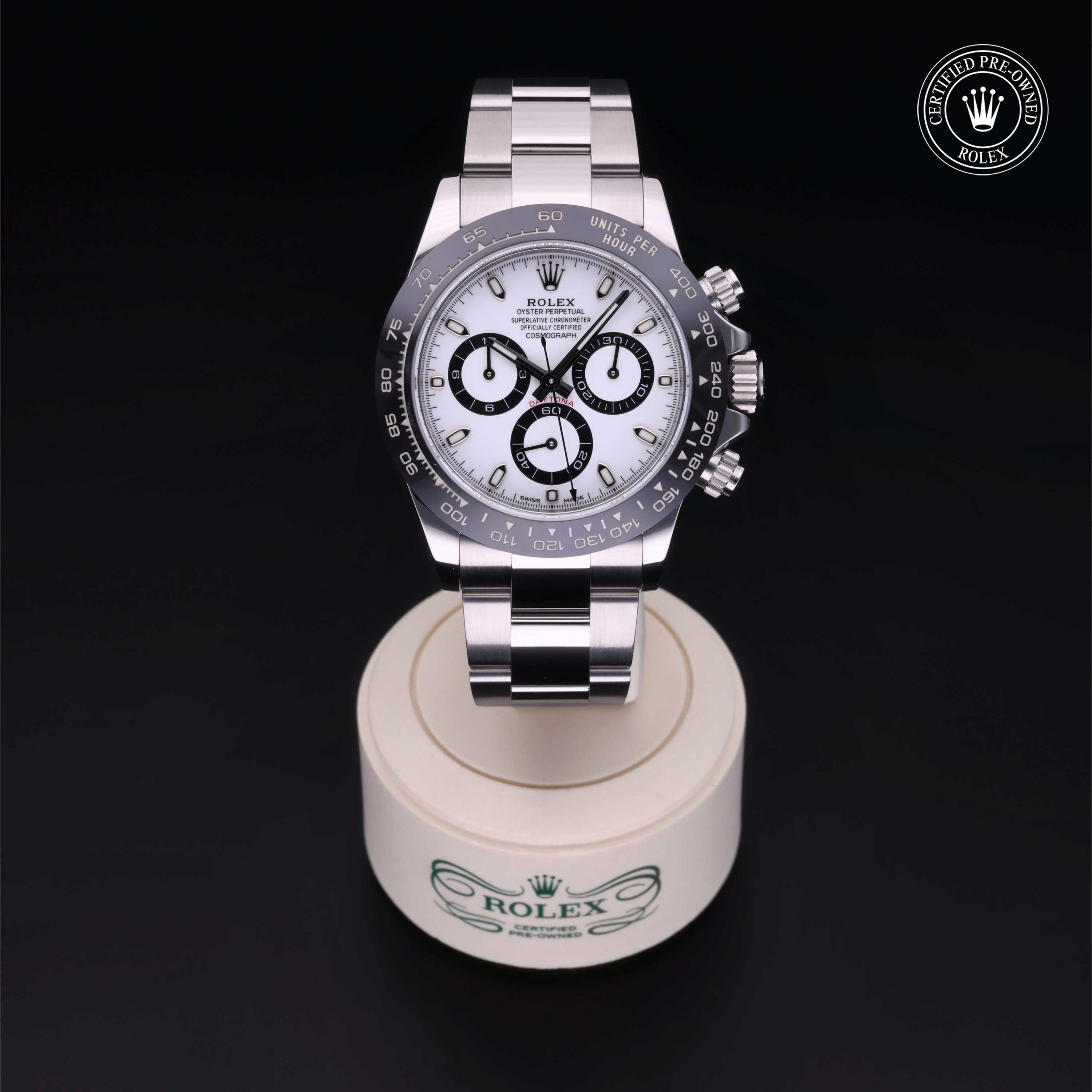 Rolex luxury watch with a stainless steel bracelet and white dial featuring black chronograph subdials, displayed on a branded stand with a certified pre-owned Rolex logo.