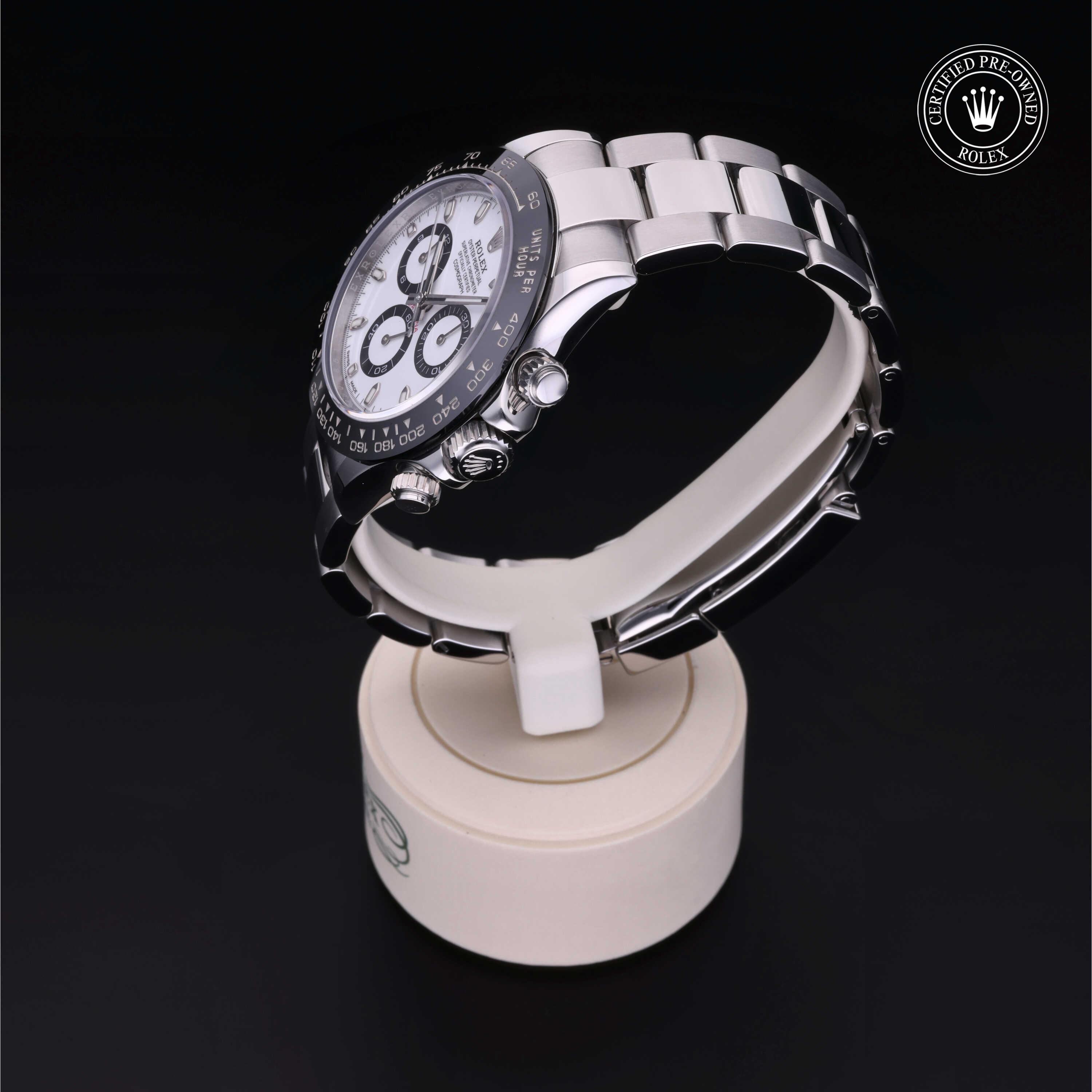Silver luxury watch with a white dial, black bezel, and chronograph features displayed on a white stand against a black background, featuring a certified pre-owned Rolex emblem.