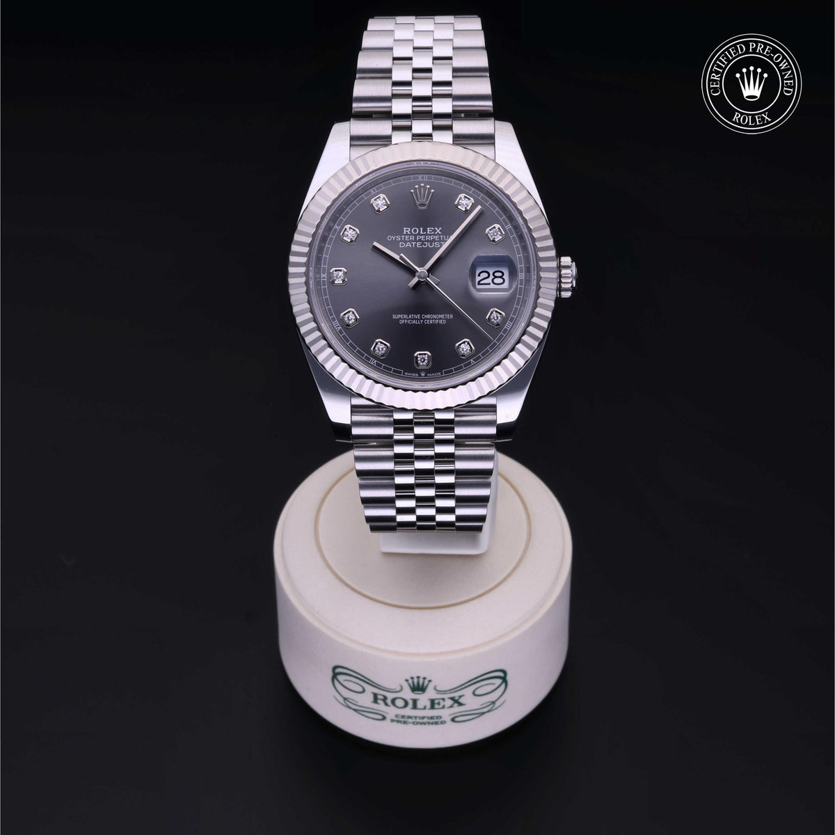 Rolex Datejust watch with silver jubilee bracelet, dark gray dial featuring diamond hour markers, displayed on a branded white stand with a certified pre-owned Rolex logo in the corner.